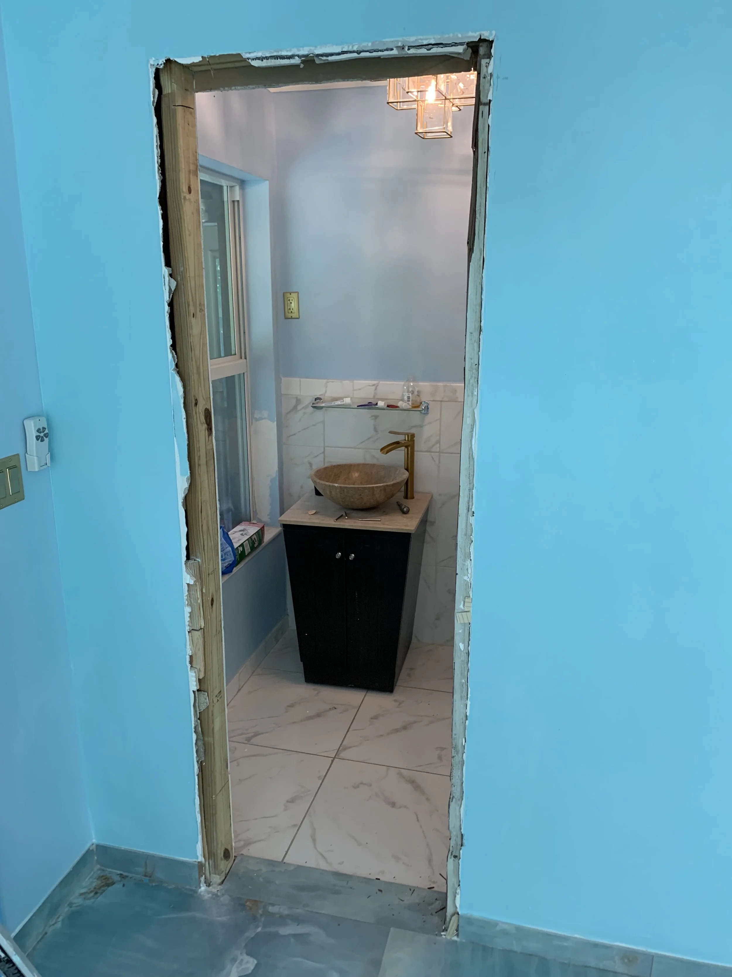 A bathroom under renovation with an unfinished doorway, showing a small black vanity with a stone vessel sink and gold faucet, marble floor and wall tiles, a window, and a modern chandelier.