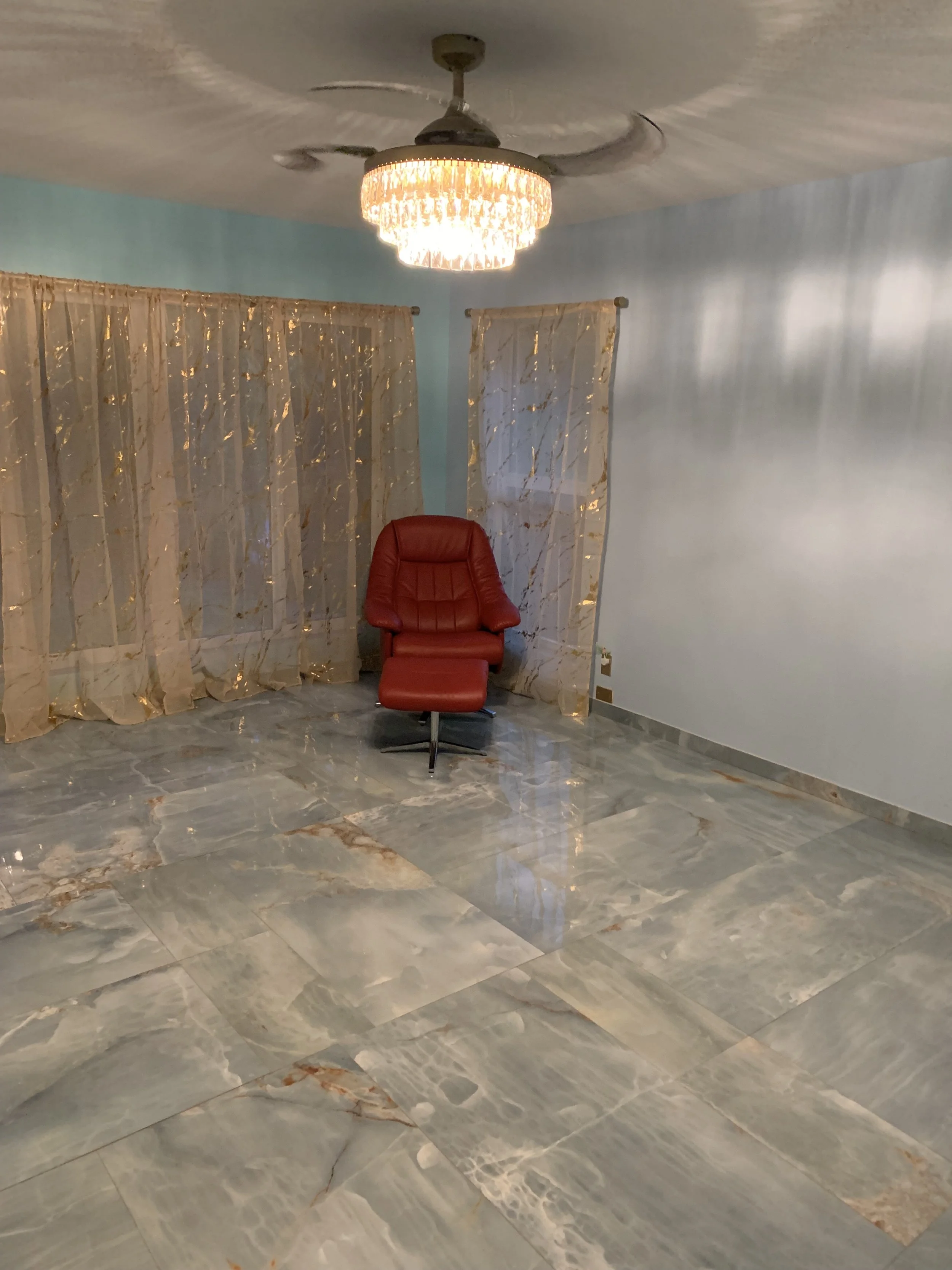 Empty room with shiny marble flooring, light blue walls, beige patterned curtains, a ceiling fan with a crystal chandelier, and a red leather chair in the corner.