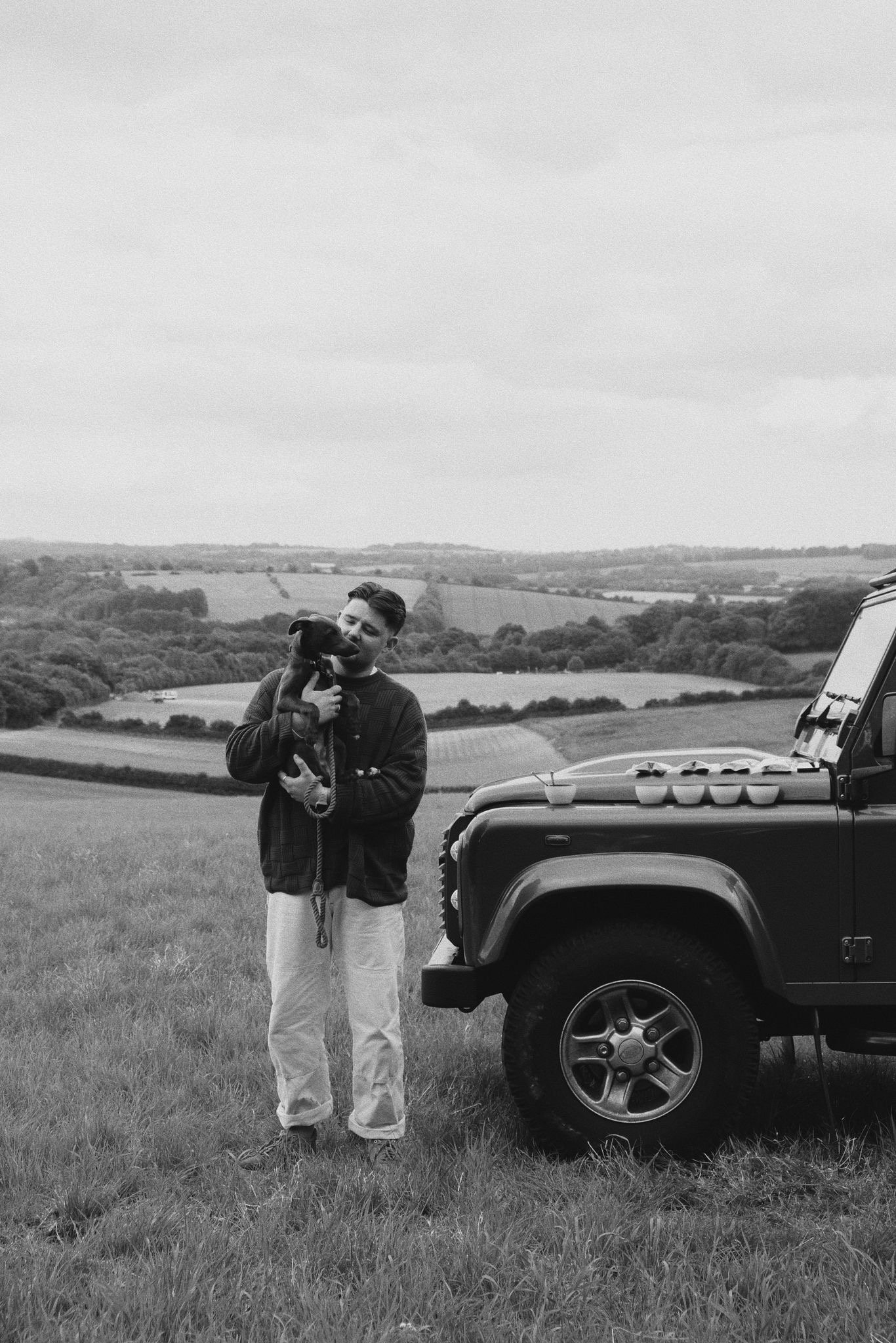 A person standing on a grassy field holding a dog, with a black off-road vehicle nearby, and rolling hills and open countryside in the background.