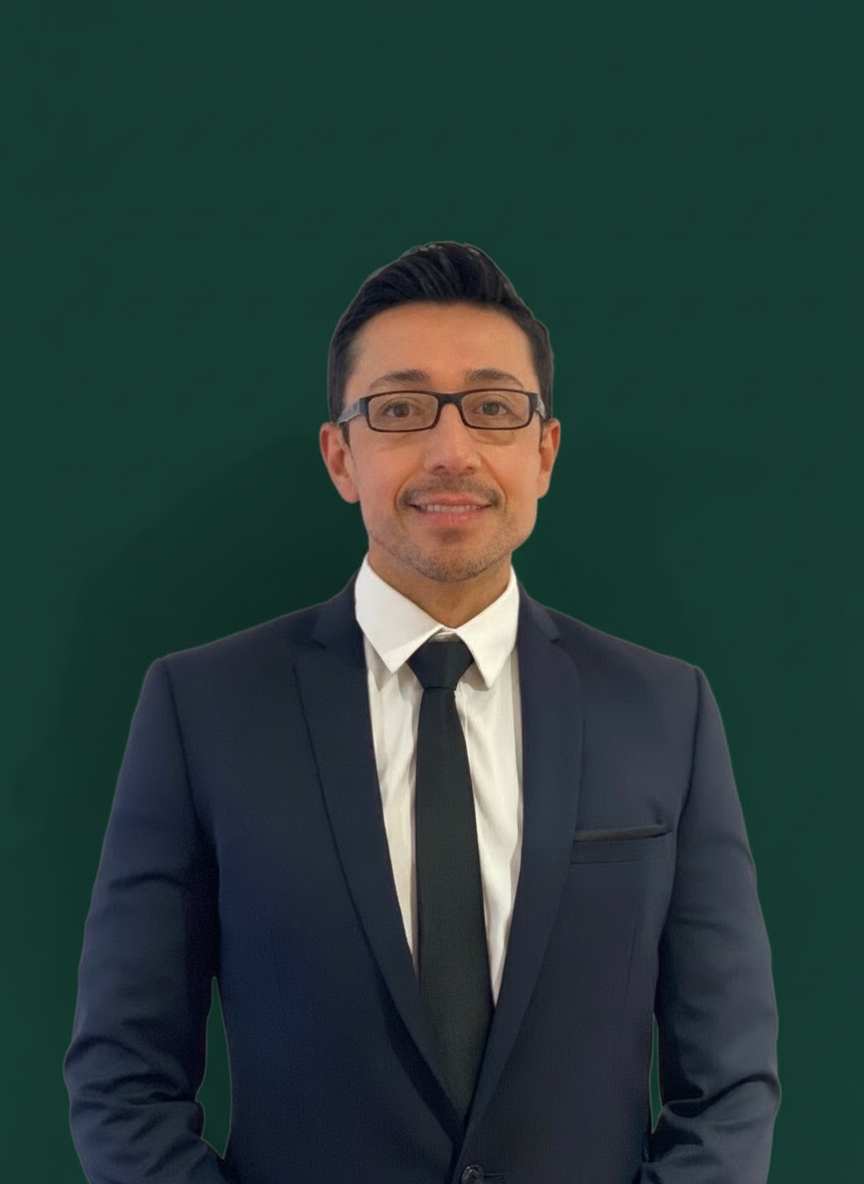 A man with dark hair and glasses wearing a dark suit, white shirt, and black tie, smiling at the camera, standing against a dark green background.