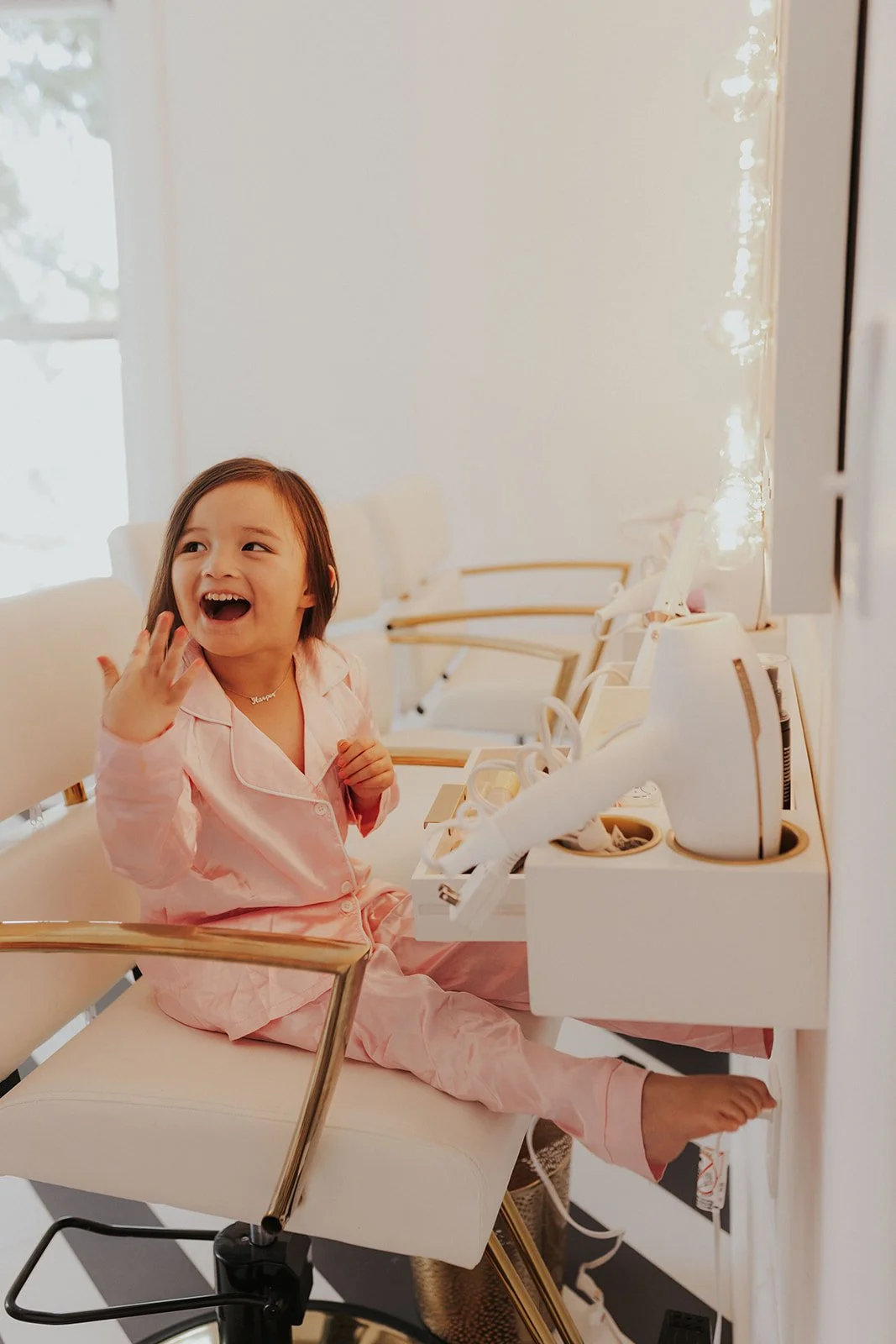 A young girl sitting at a skincare or makeup station, smiling and waving, wearing pink pajamas, with a mirror and beauty products in front of her.