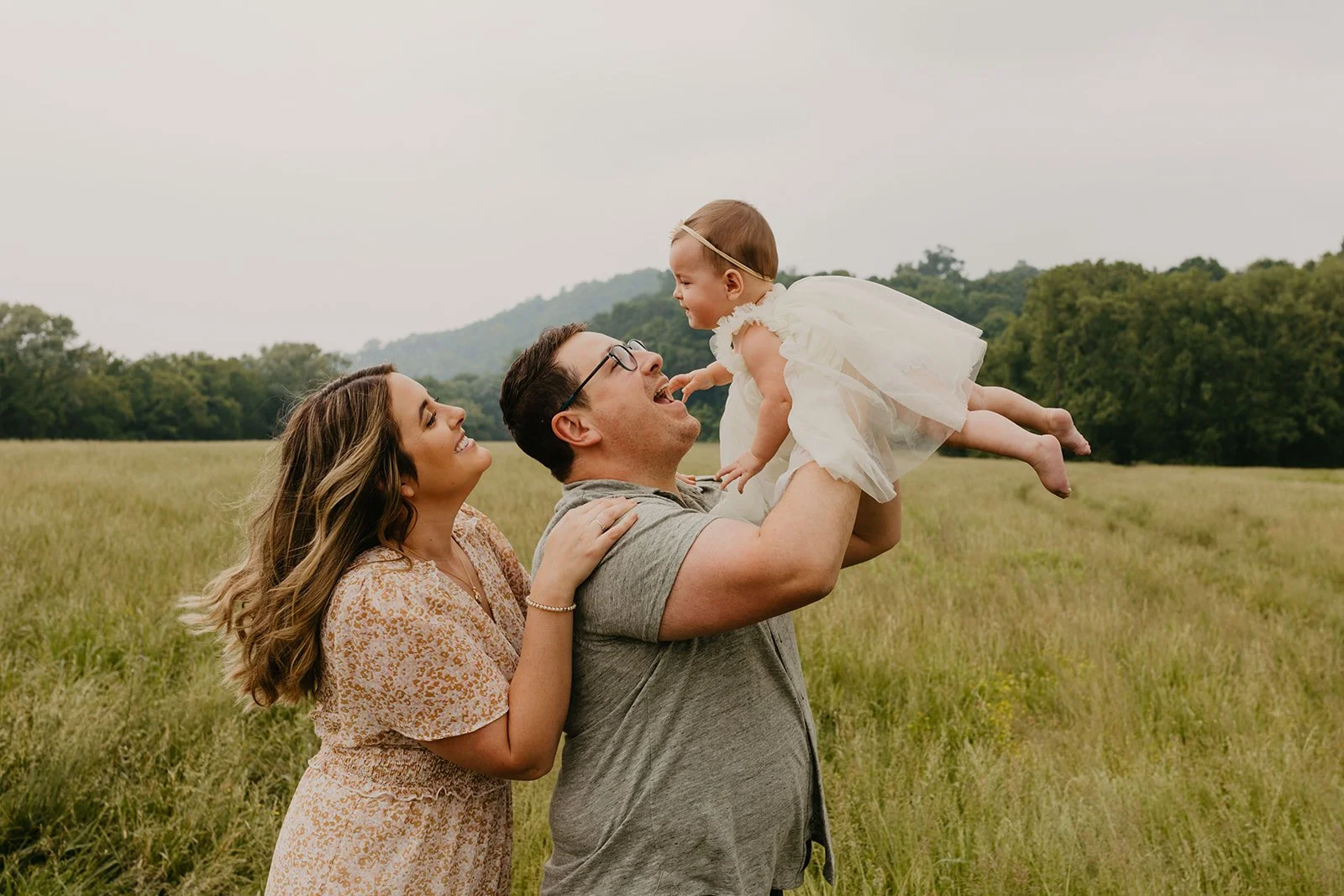Family of three in a field, with a man holding a young girl in a white dress and the woman smiling, all enjoying an outdoor moment.