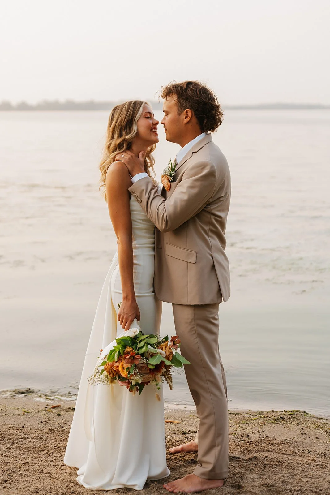A bride and groom are standing close by the water at sunset, touching foreheads and smiling. The bride holds a bouquet of flowers, and the groom is barefoot, wearing a beige suit and a boutonniere.