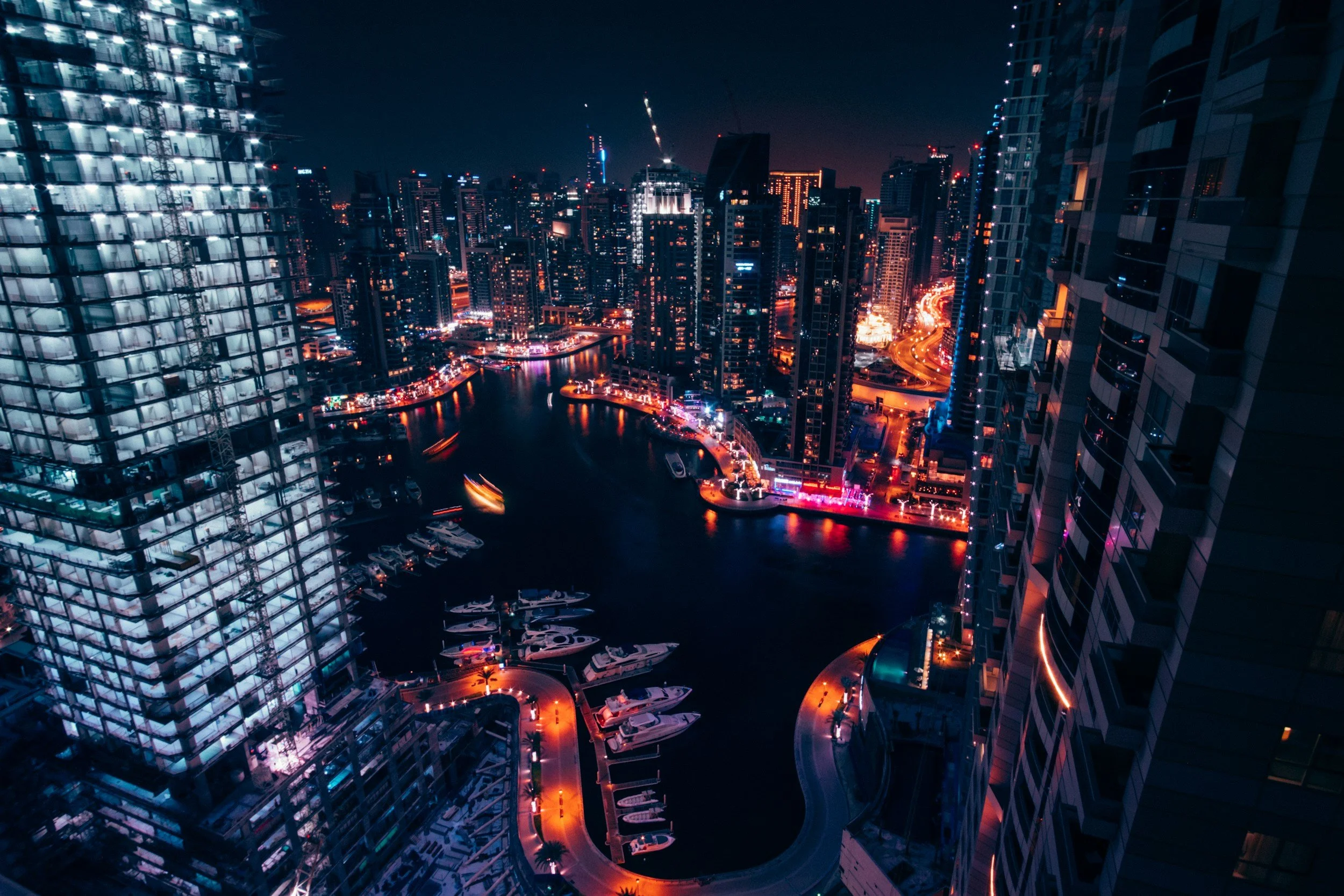 Nighttime cityscape with illuminated skyscrapers, a marina with boats, and winding roads, viewed from a high vantage point.