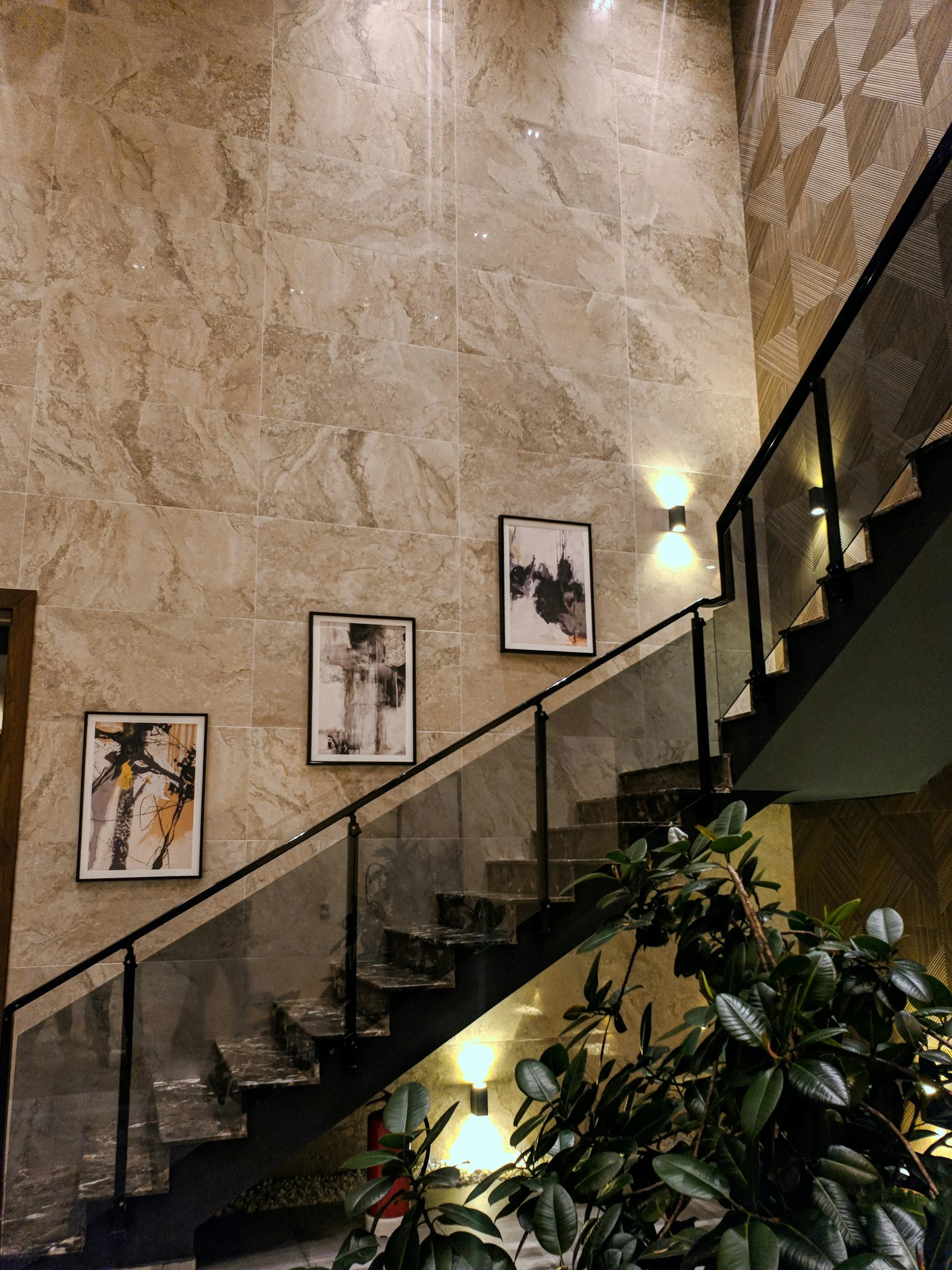 Interior view of a staircase with black marble steps and a glass railing, beige marble wall with three framed abstract art pieces, green leafy plant near the bottom of the staircase, and warm wall-mounted lighting.