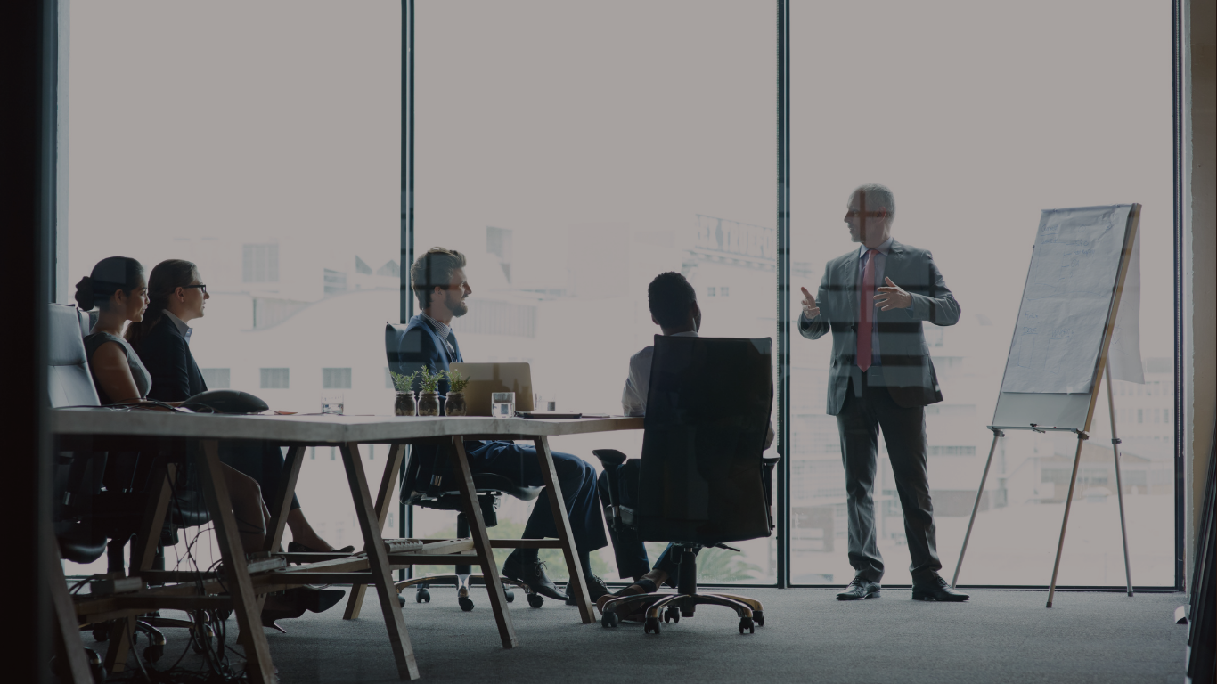 A businessman in a suit giving a presentation to four colleagues in a conference room with large windows.