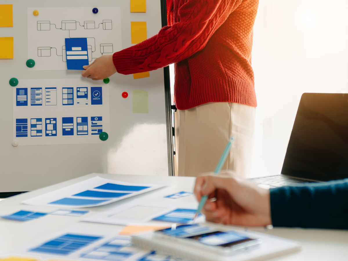 Person in a red sweater pointing at a flowchart on a whiteboard, with a person nearby taking notes and a laptop on the table.