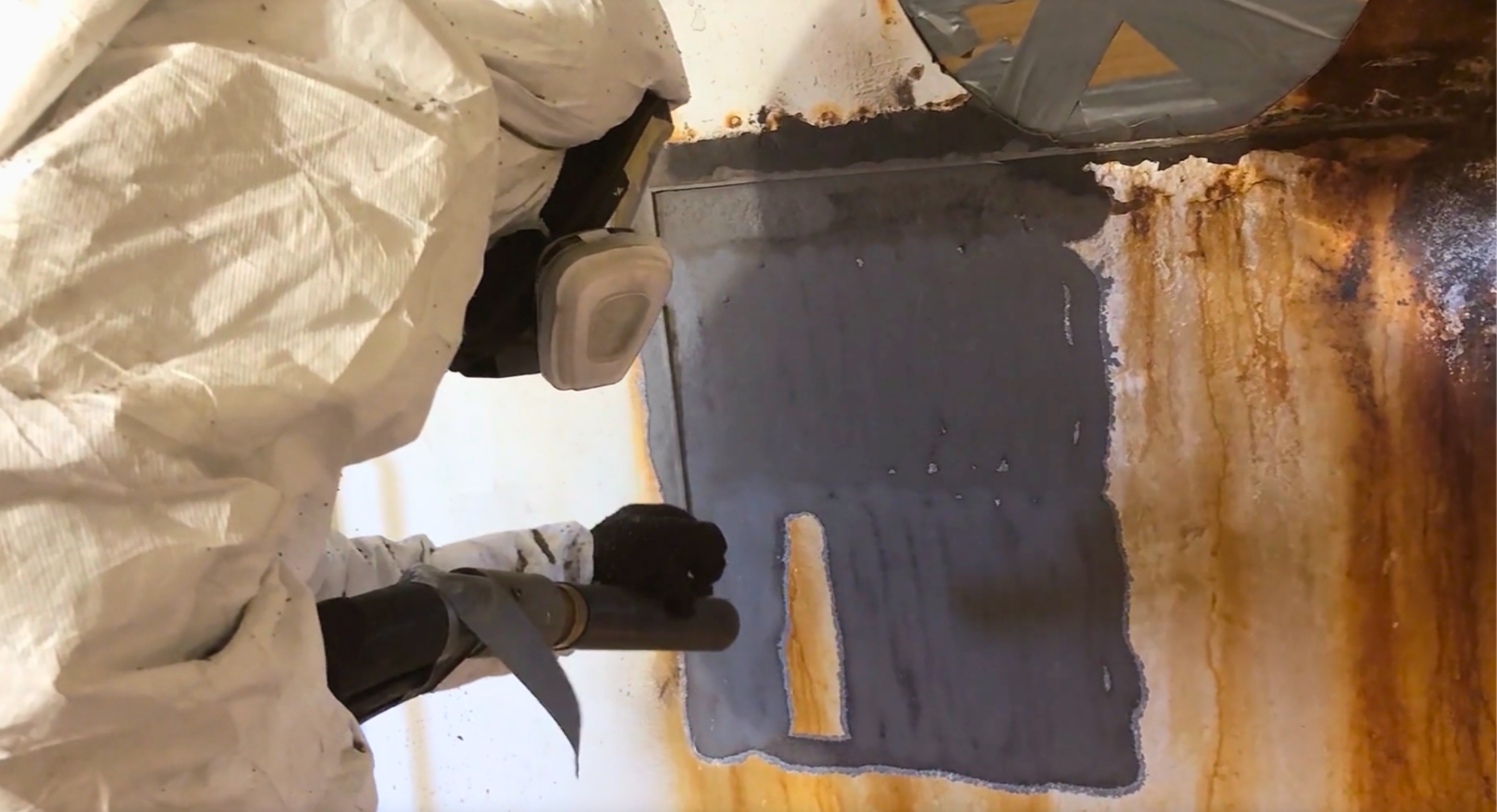 Person in protective clothing using a pipe to scrape and clean rust and corrosion from a metal surface with rust stains and corrosion residues.