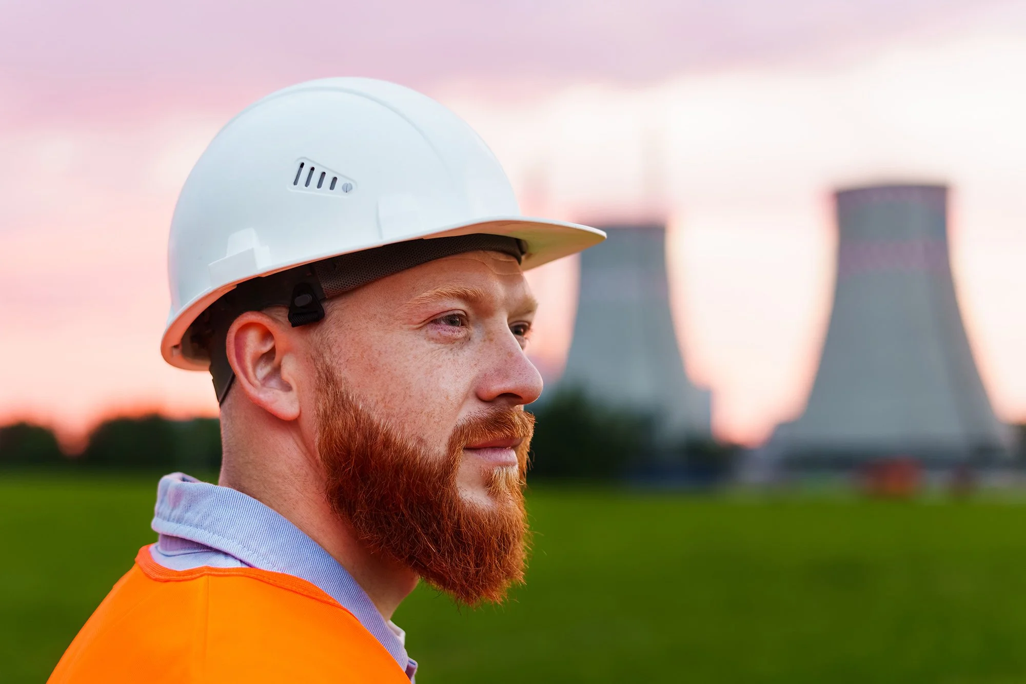 A man with red hair and a beard wearing a white safety helmet and an orange safety vest at a construction site with cooling towers in the background during sunset.