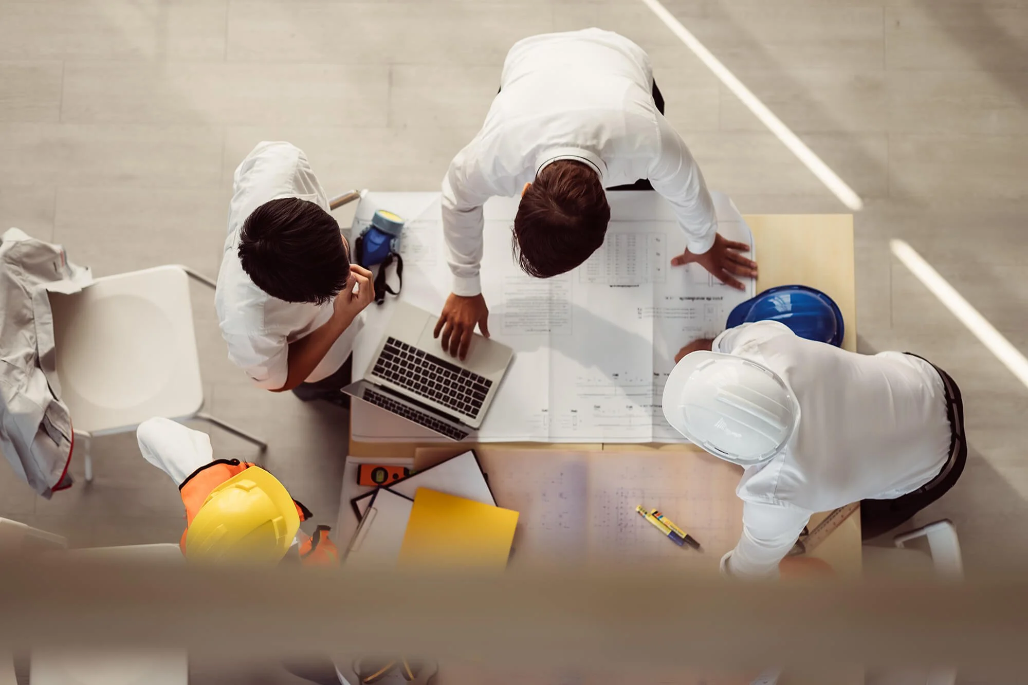 Overhead view of four engineers and construction workers around a table discussing blueprints and plans, with laptops, helmets, and construction tools on the table.