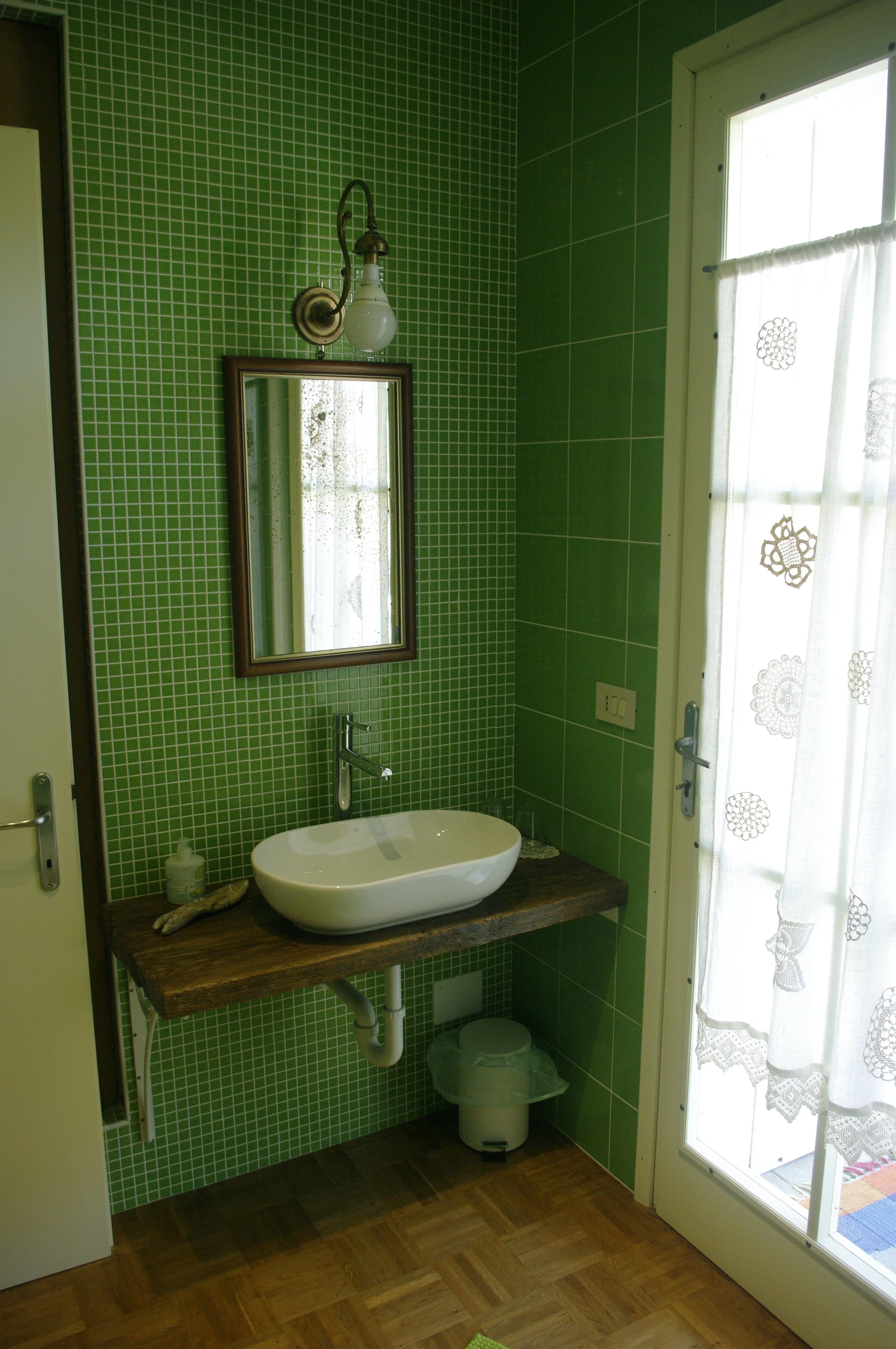 Bagno con pareti verdi, piccolo lavabo rotondo in pietra su mensile di legno, specchio rettangolare sopra il lavabo, lampada da parete con bulb in vetro, porta-finestra con tenda bianca e motivi decorativi, pavimento in parquet
