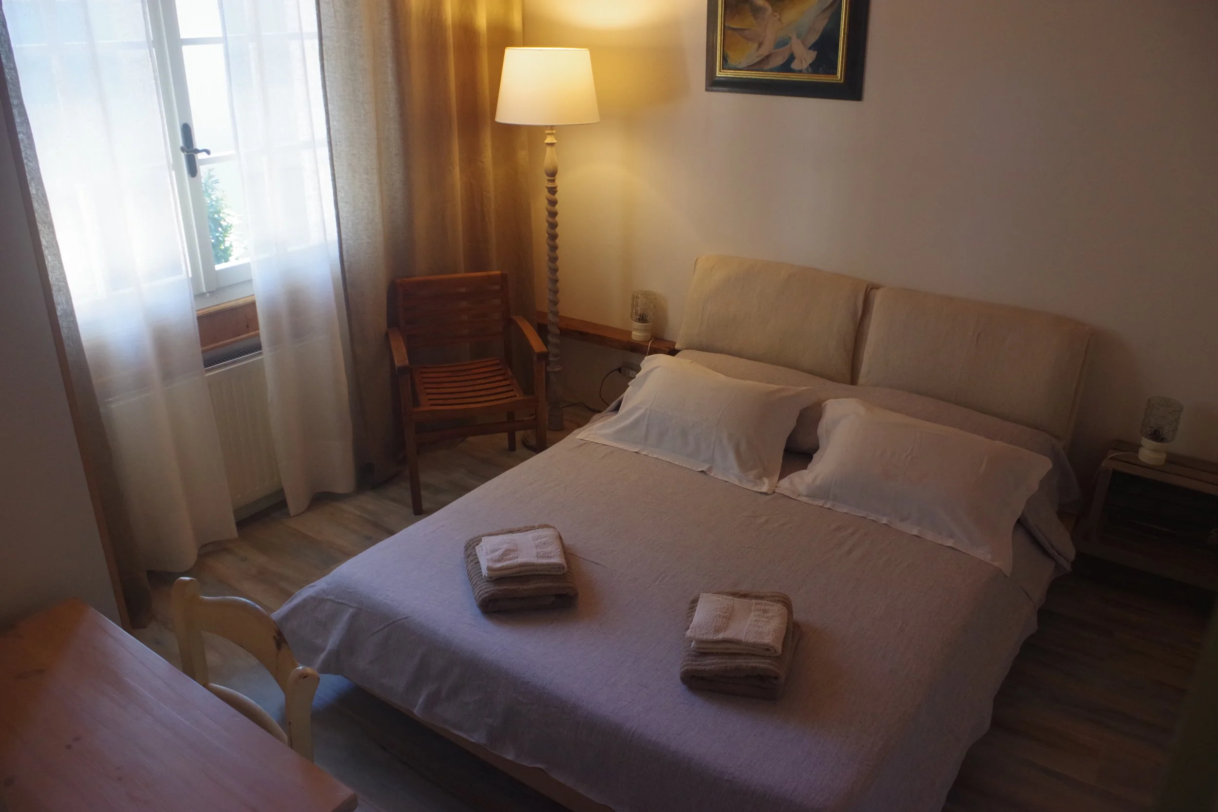 Camera da letto con un letto matrimoniale, due asciugamani piegati sul letto, una lampada da terra, una sedia di legno, tende trasparenti e un quadro appeso sulla parete.