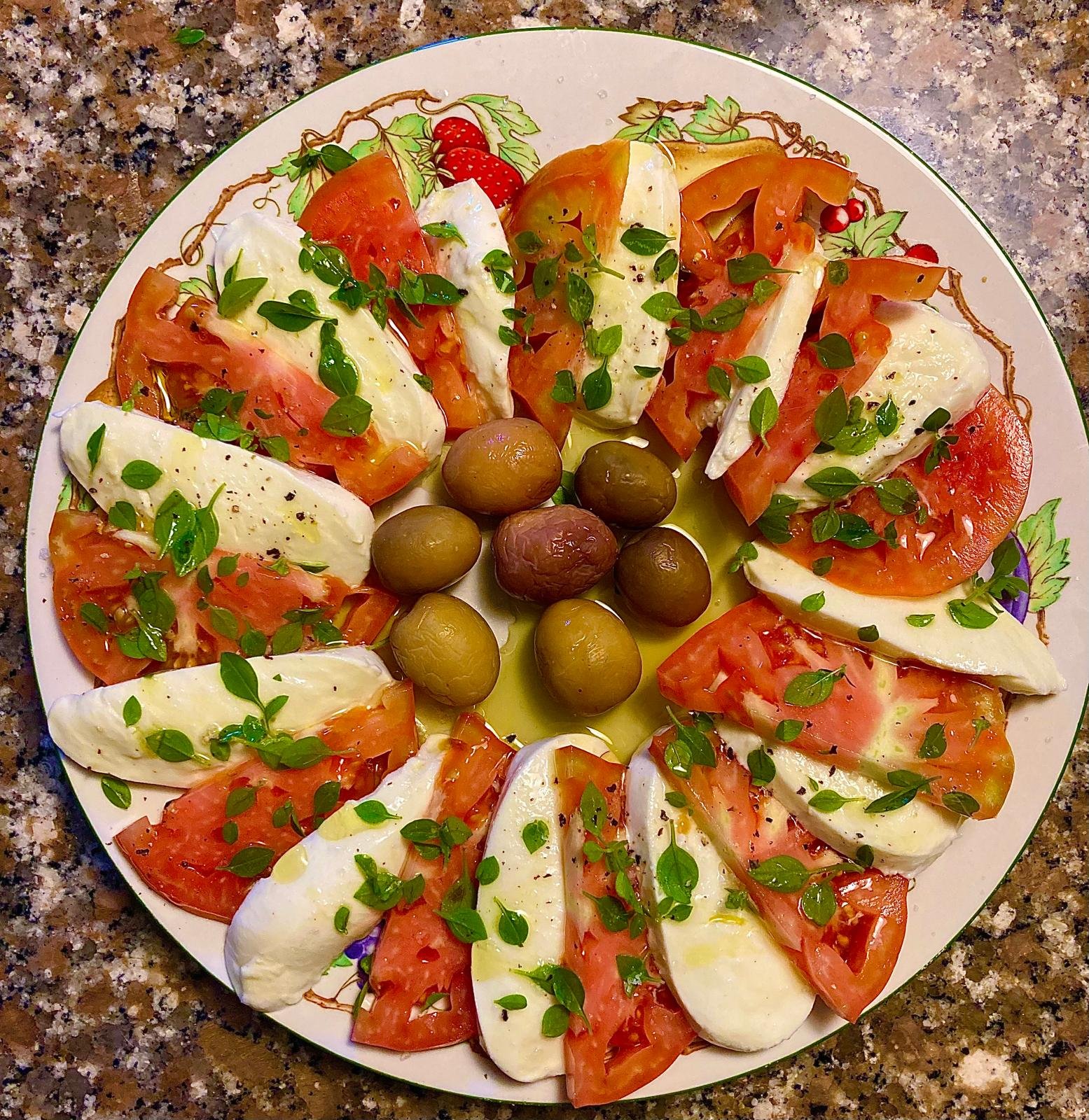 Insalata caprese con pomodori, mozzarella, olive verdi, olive viola e foglie di basilico su un piatto bianco