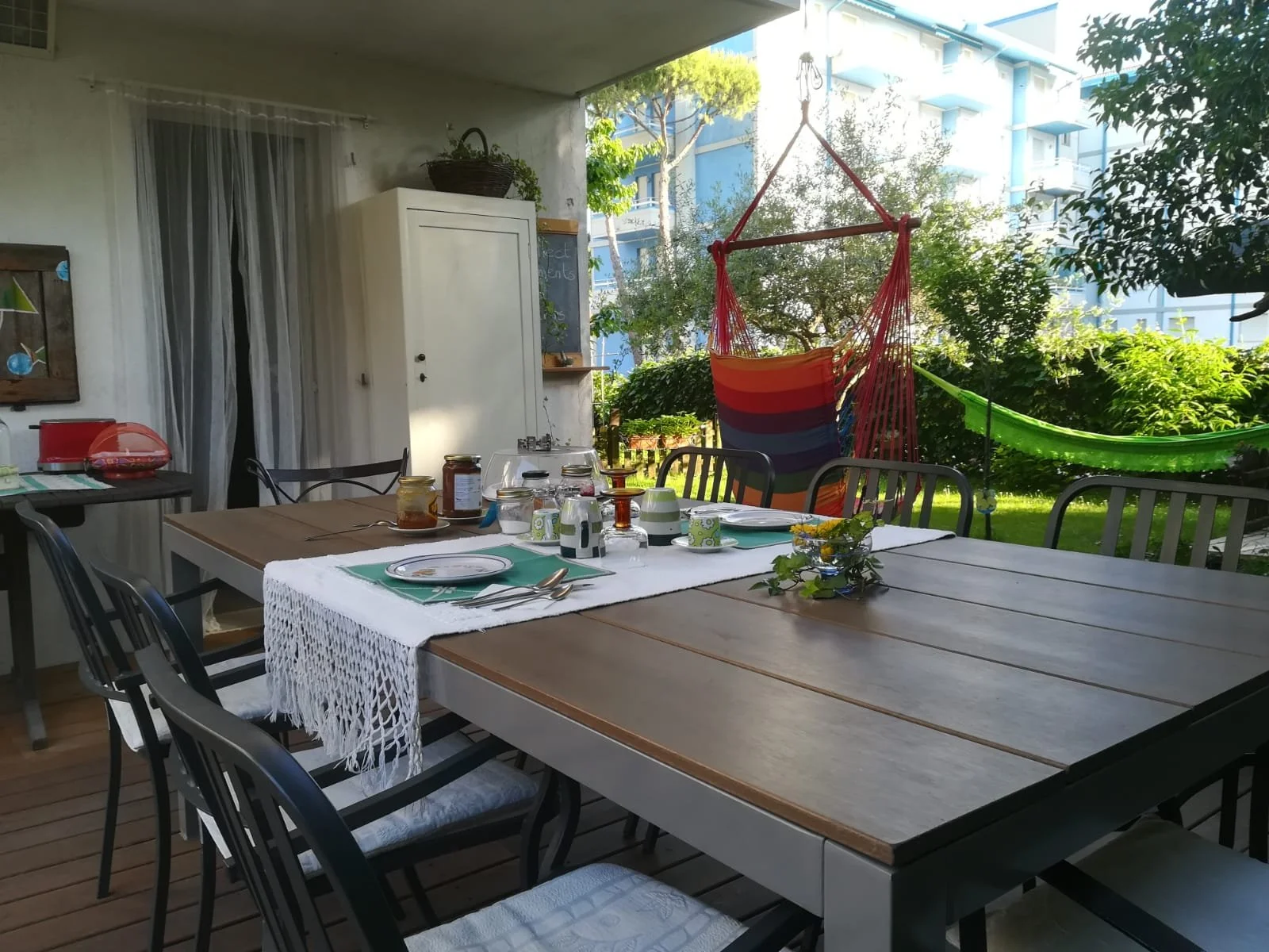 Tavolo apparecchiato per la colazione o il pranzo sotto un terrazzo con sedie nere, un'ampia libreria bianca e due amache colorate all'aperto in giardino con alberi e un edificio di moderni appartamenti sullo sfondo.