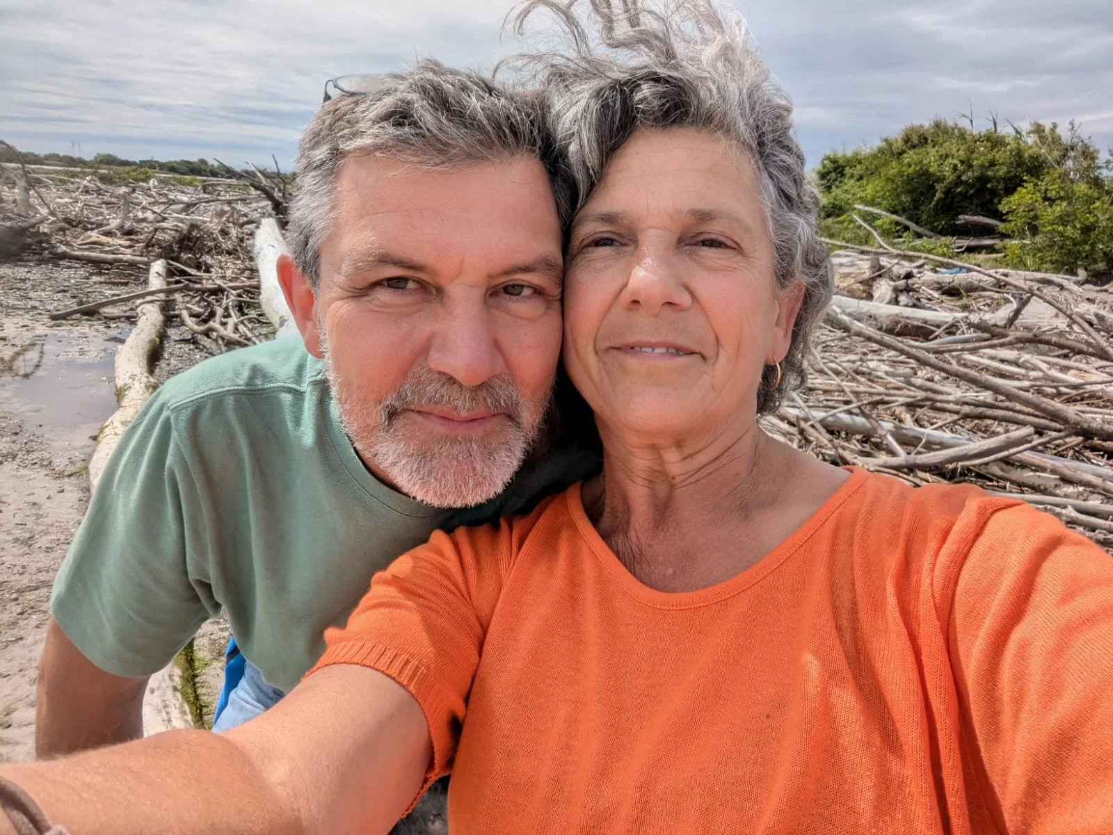 Una coppia di anziani che si abbraccia sulla spiaggia con tronchi e rami spiaggiati sullo sfondo.