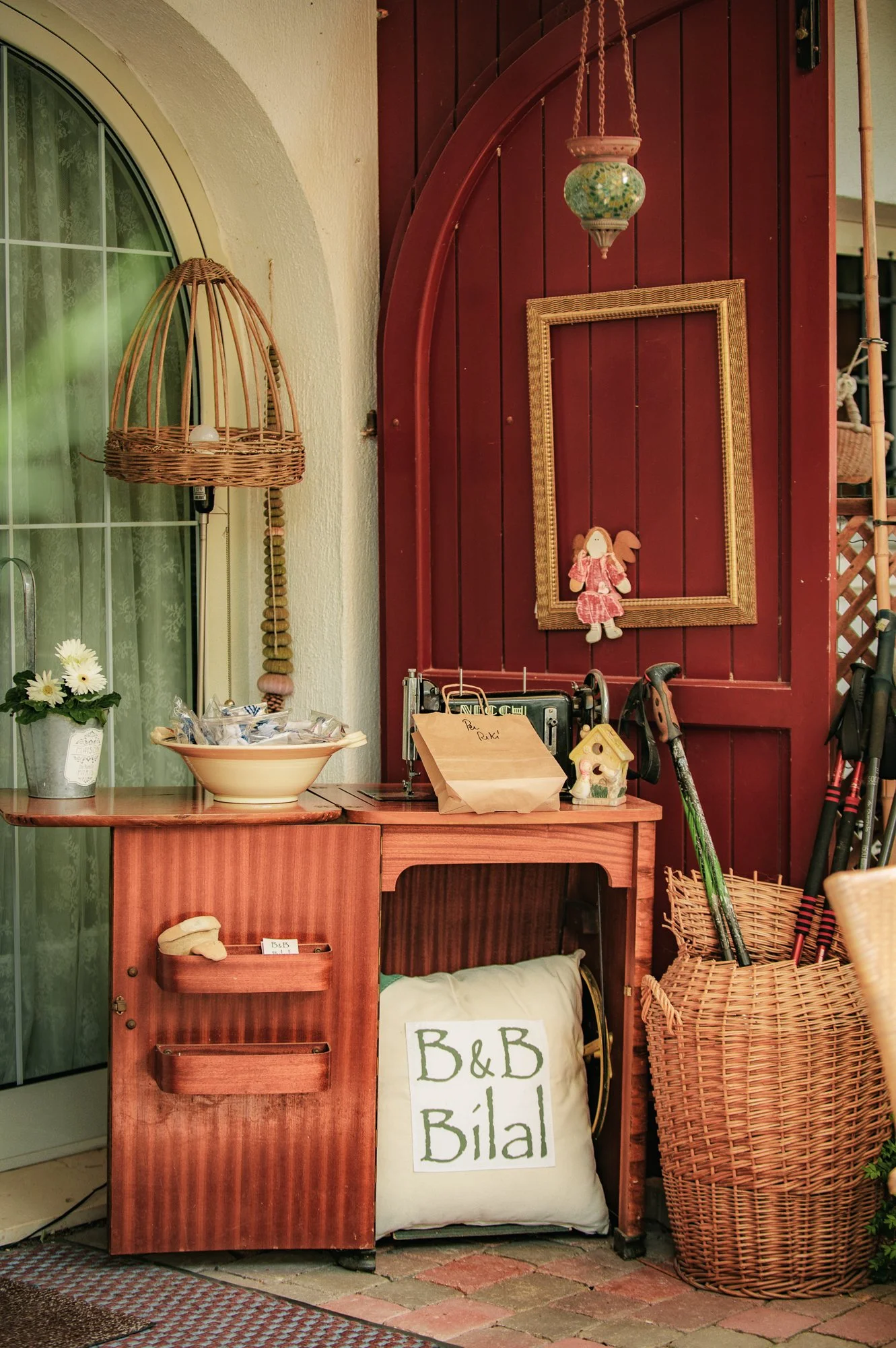 Spazio esterno con tavolo in legno, cestino di vimini con scope e bastoni, cornice vuota su pannello di legno rosso, decorazioni come bambola, lampada in vimini, vaso con fiori, borsa con scritto 'B&B Bilal' e vari oggetti.
