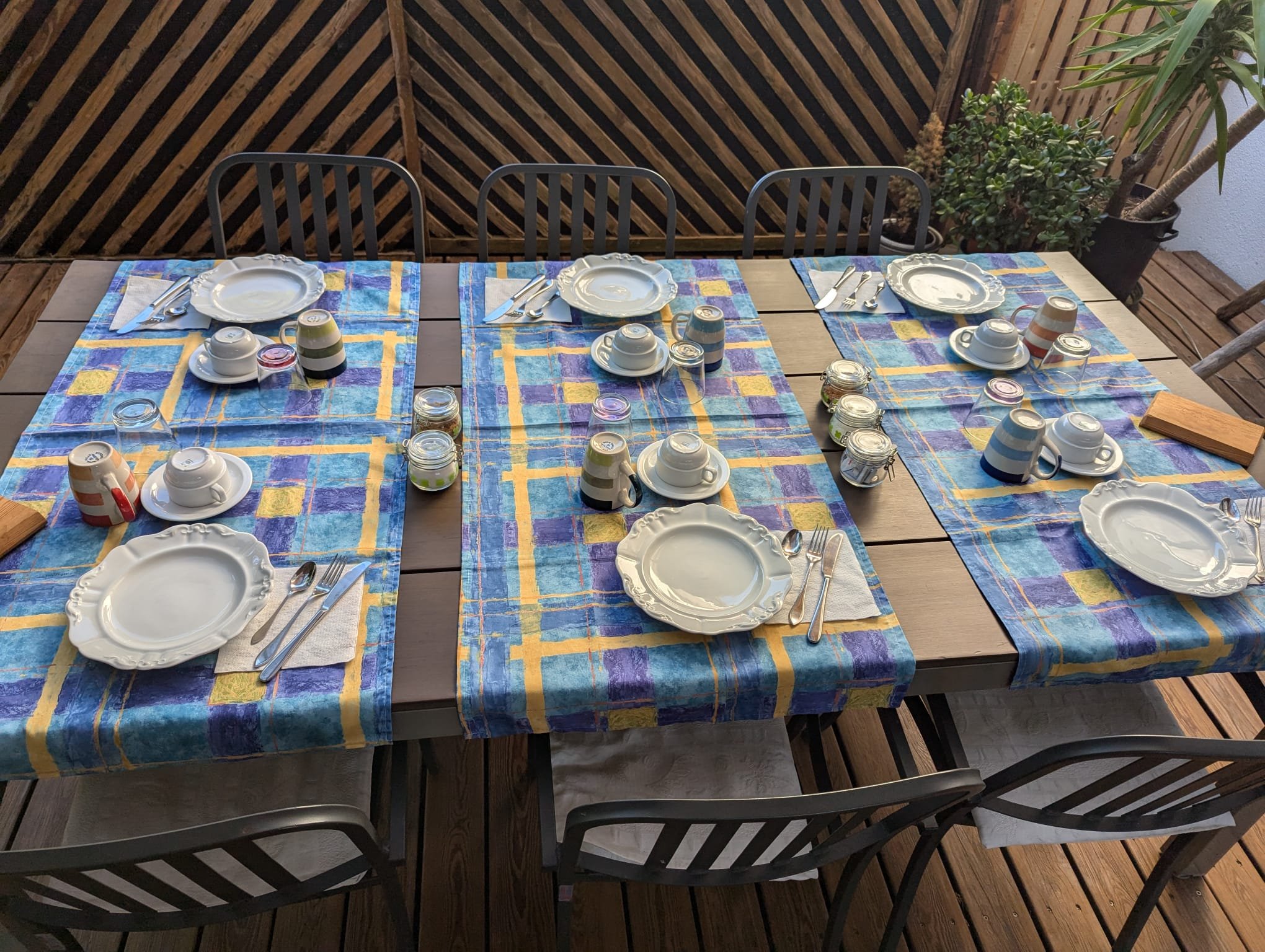 Tavolo da pranzo all'aperto apparecchiato con piatti bianchi decorativi, bicchieri, tazzine con piattini, posate, ciotole con coperchio, e tessuti a motivo colorato, in un'area con piante e parete in bambù.