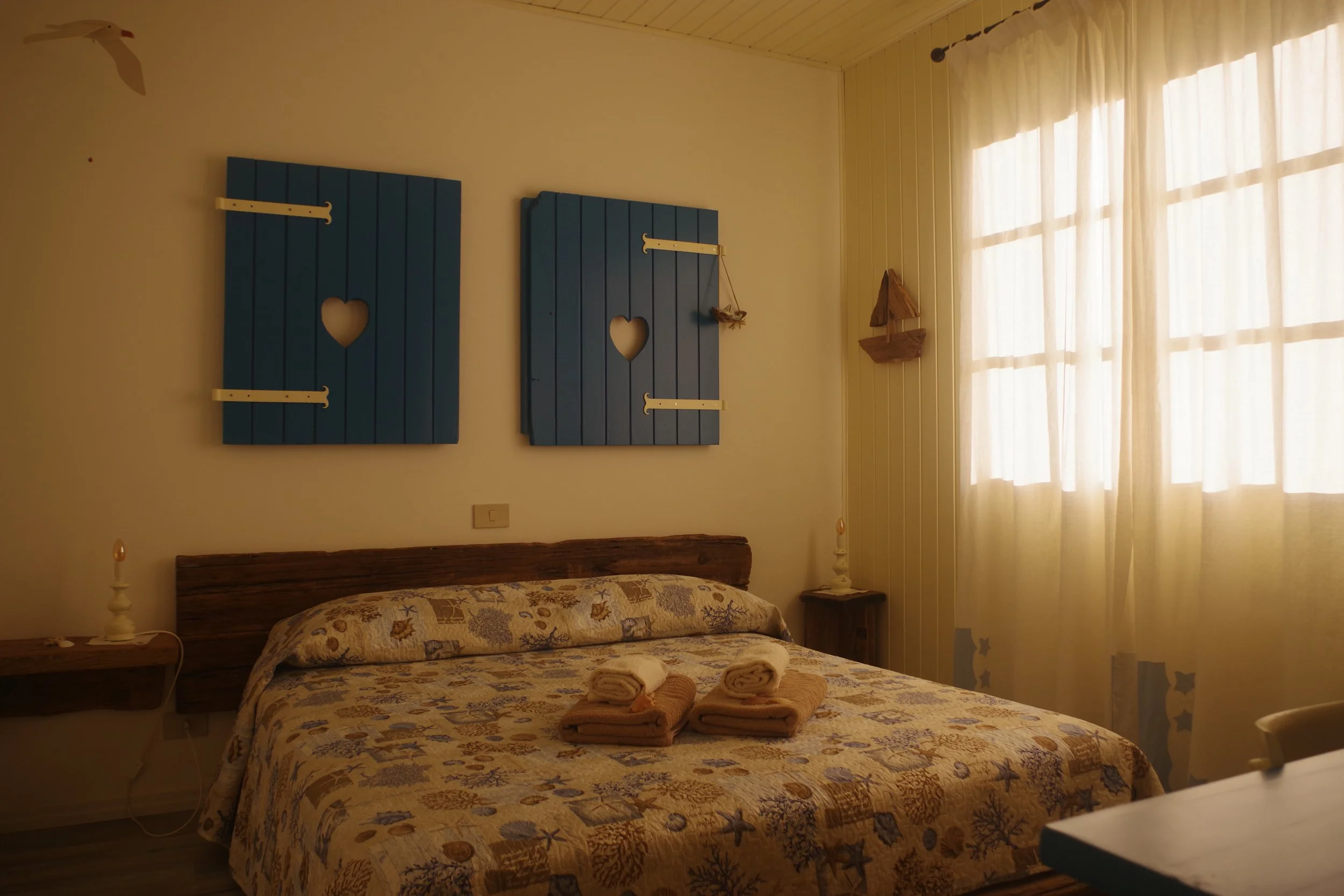 Stanza con letto matrimoniale, tende leggere, pareti bianche, due quadri di legno blu con cuori e cerniere, lampade accanto al letto, asciugamani sulla coperta, decorazioni in stile marino.