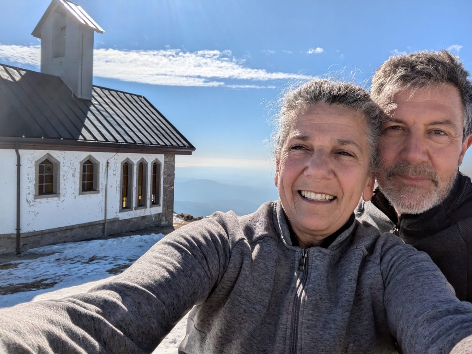 Una coppia di due persone anziane, una donna e un uomo, sorridenti e che fanno un selfie in montagna davanti a una piccola chiesa con tetto in metallo e pareti bianche, con il cielo blu e nuvole leggere sullo sfondo.