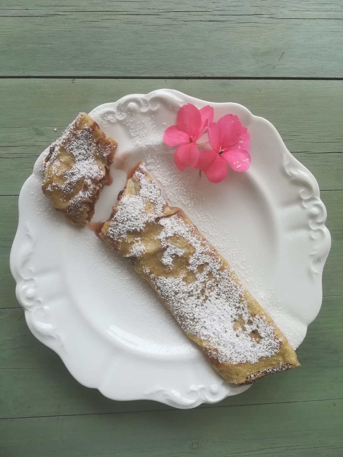 Dolce con due pezzi di torta rivestiti di zucchero a velo, decorato con fiori rosa, su piatto bianco con bordo decorato, sopra tavolo verde.