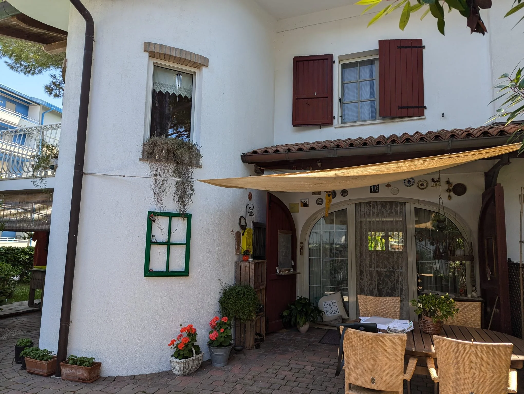 Esterno di una casa con pareti bianche, persiane rosse, una finestra, un portico con arredi, piante e fiori.