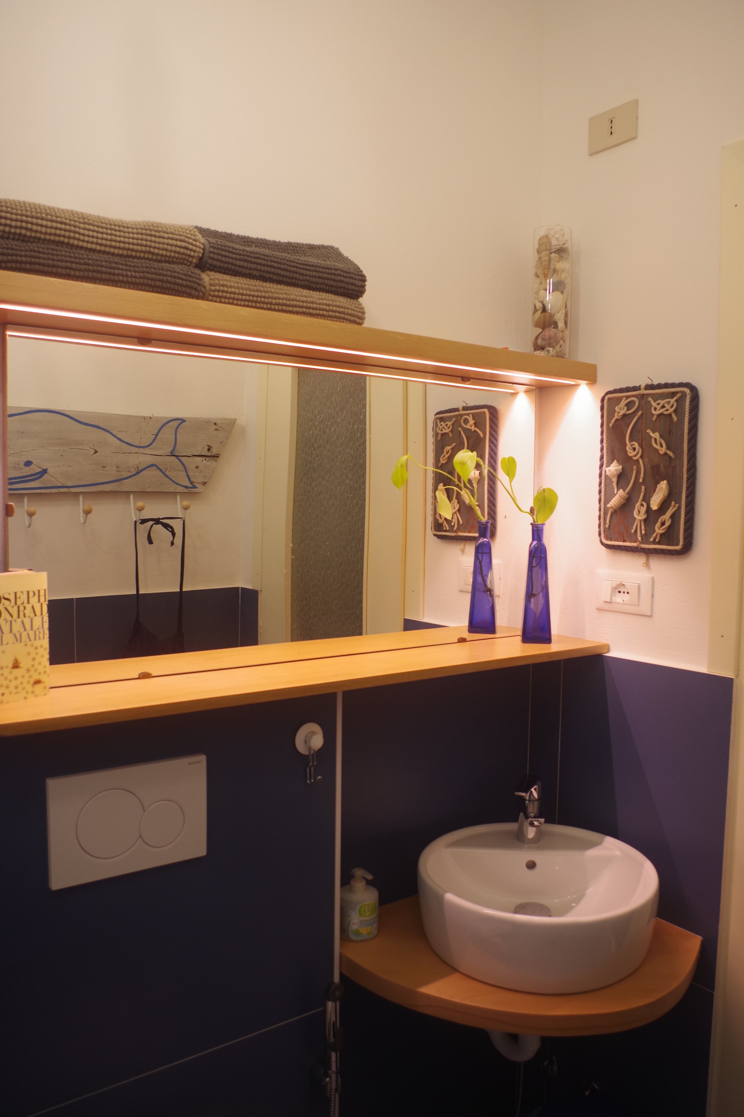 Questo è un interno di un bagno con lavabo rotondo di ceramica, due bottiglie blu con piante verdi, quadri con argilla modellata, una set di asciugamani sopra il bar e un disegno di un pesce in legno sulla parete.
