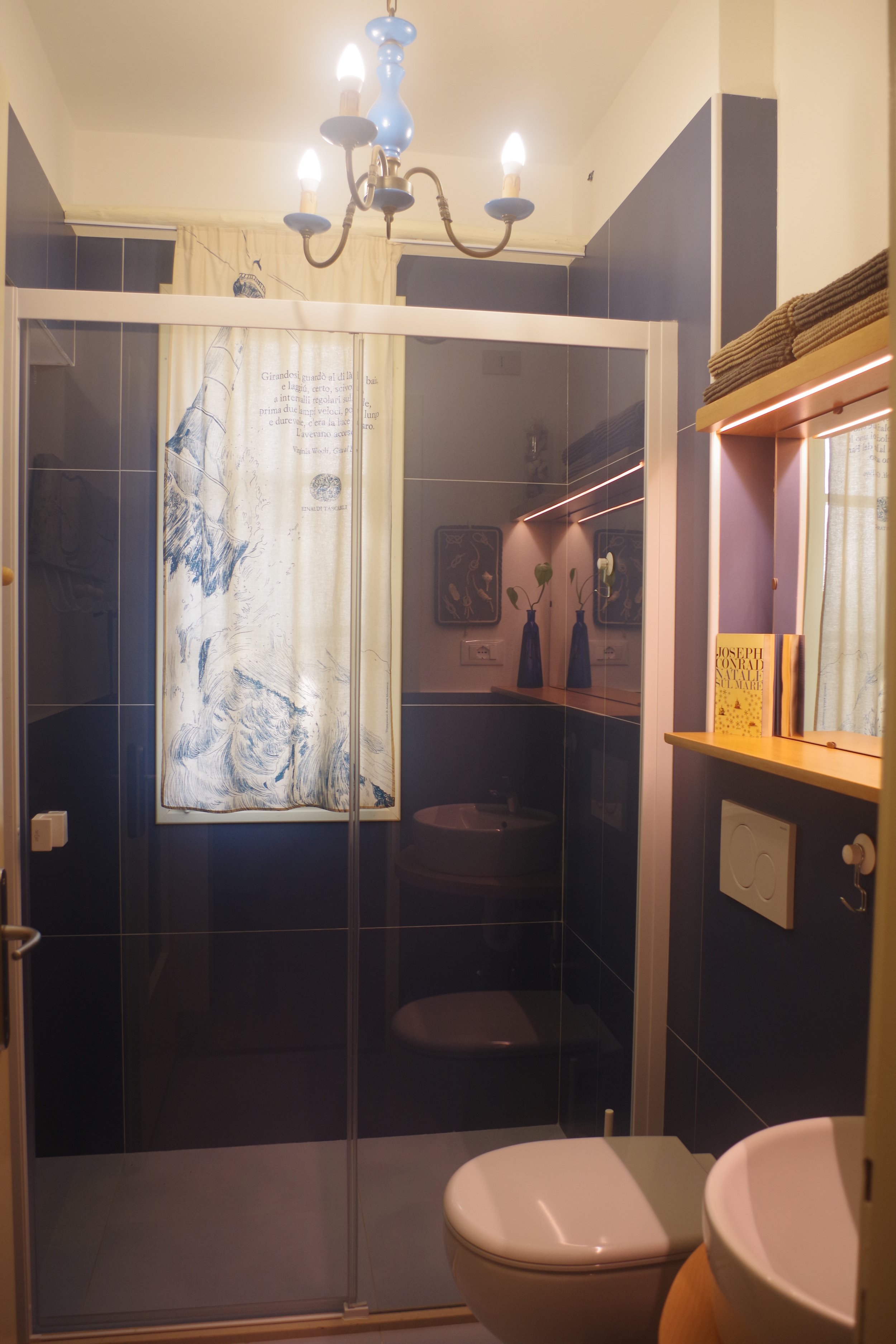 Bagno con pareti nere con piastrelle lucide, doccia con porta a scorrimento, finestra con tenda chiara con disegno, speccho con lampada, scaffale con asciugamani e libro.