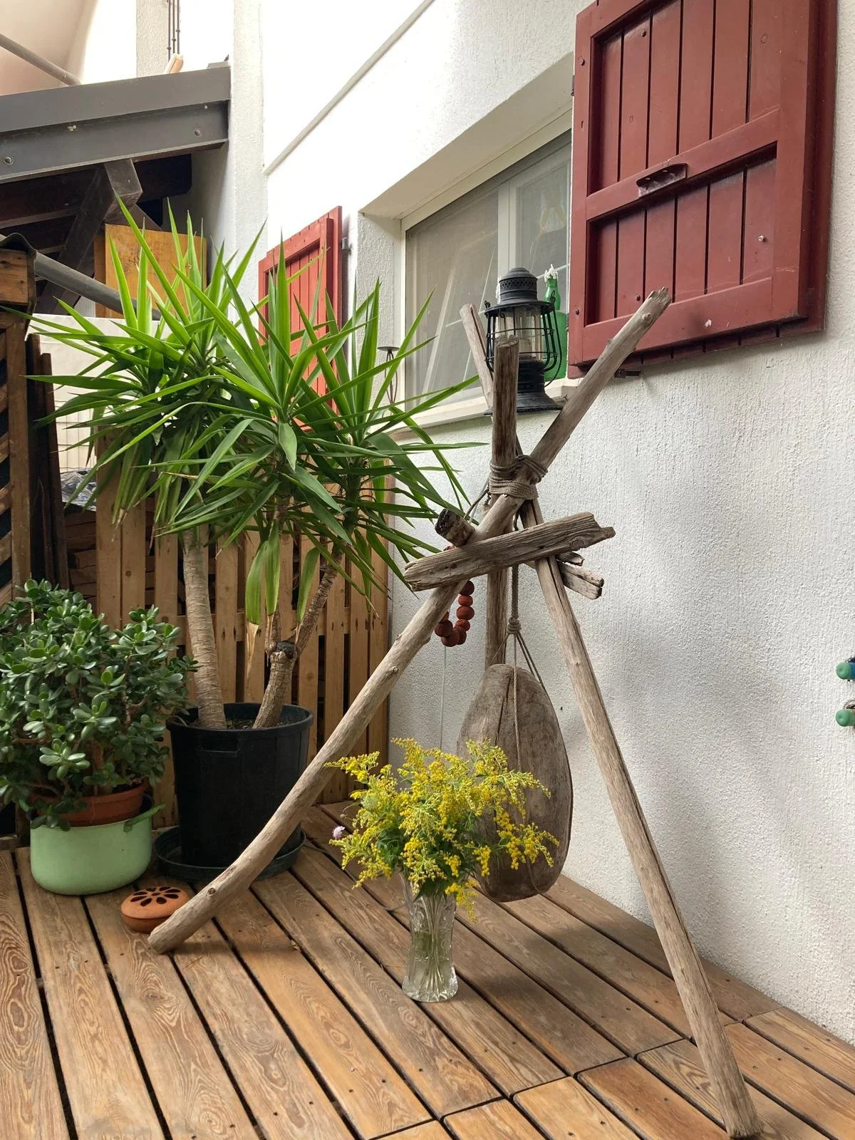 Balcone con piante in vaso, una lanterna e decorazioni, con finestra con tapparelle rosse e parete bianca.