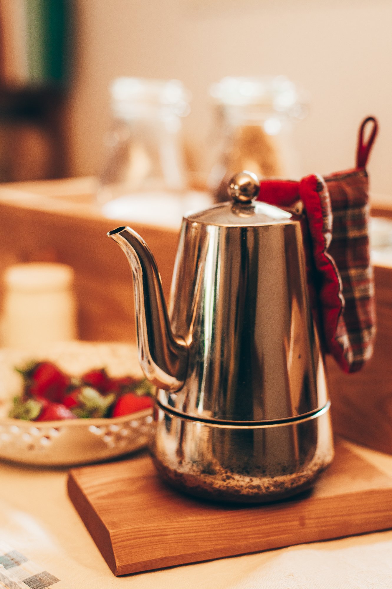 Una teiera d'acciaio lucido con un panno appoggiato sul manico, posizionata su un tagliere di legno, con una ciotola di fragole sfocate sullo sfondo.