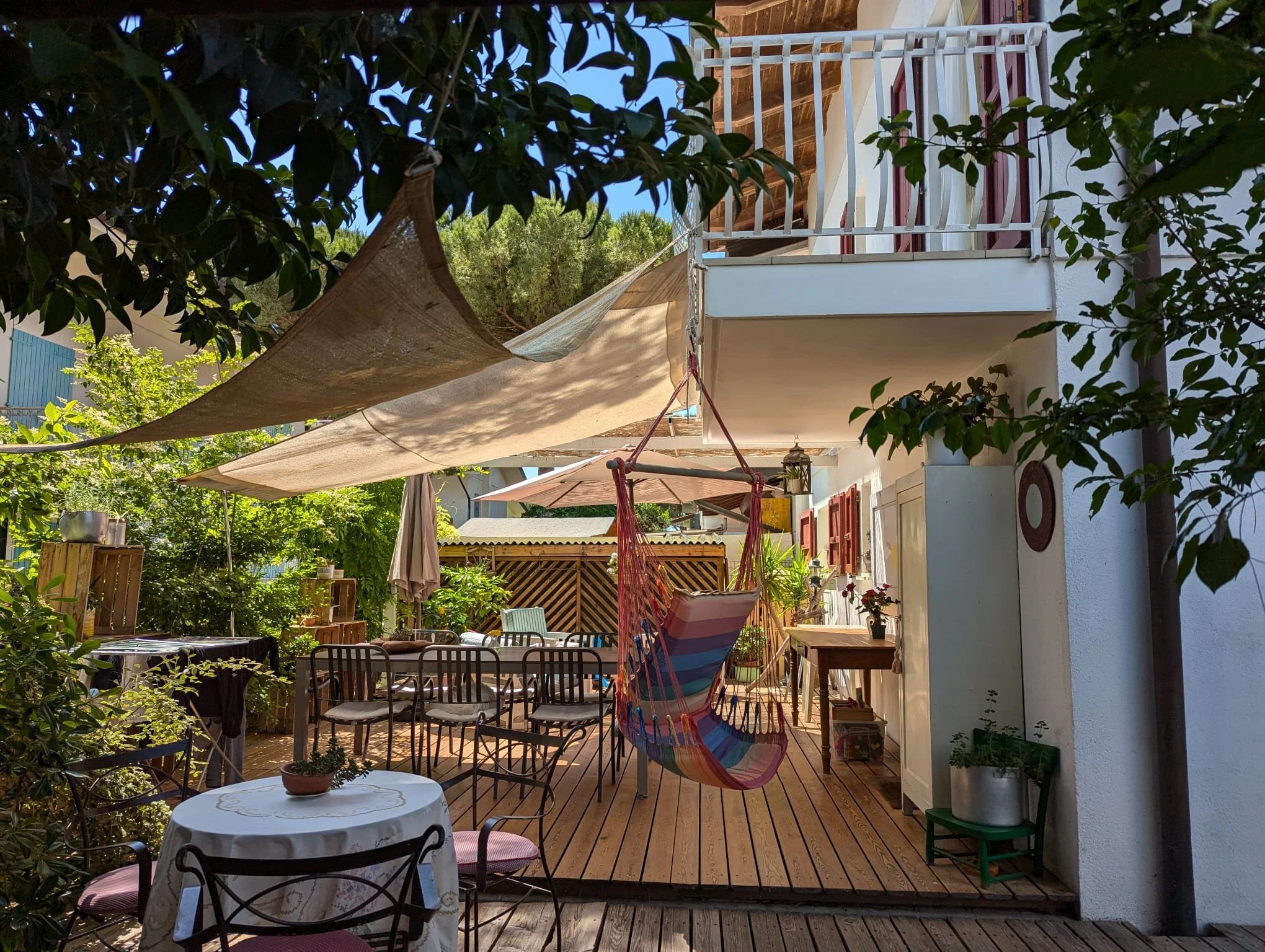 Una terrazza con tavoli, sedie, un'amaca colorata, ombrelloni, piante e decorazioni, sotto un grande ombrello beige e un tettuccio di tessuto beige.