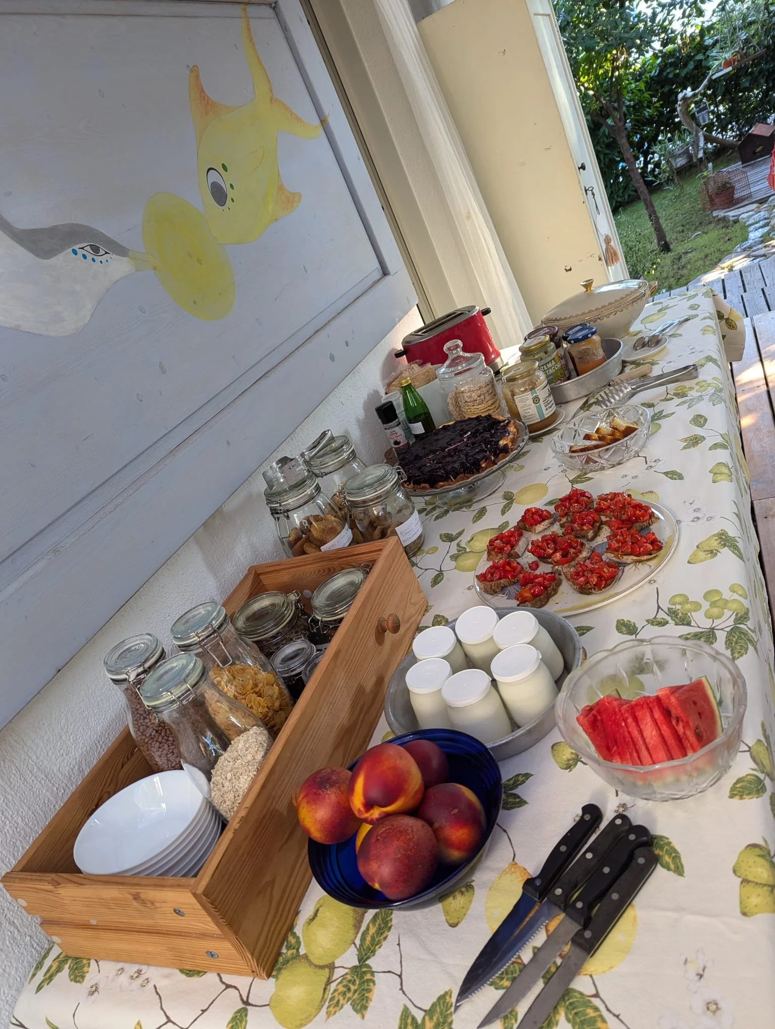 Tavolo apparecchiato con frutta, marmellate, yogurt e vari dolci, in una cucina con vista sul giardino.