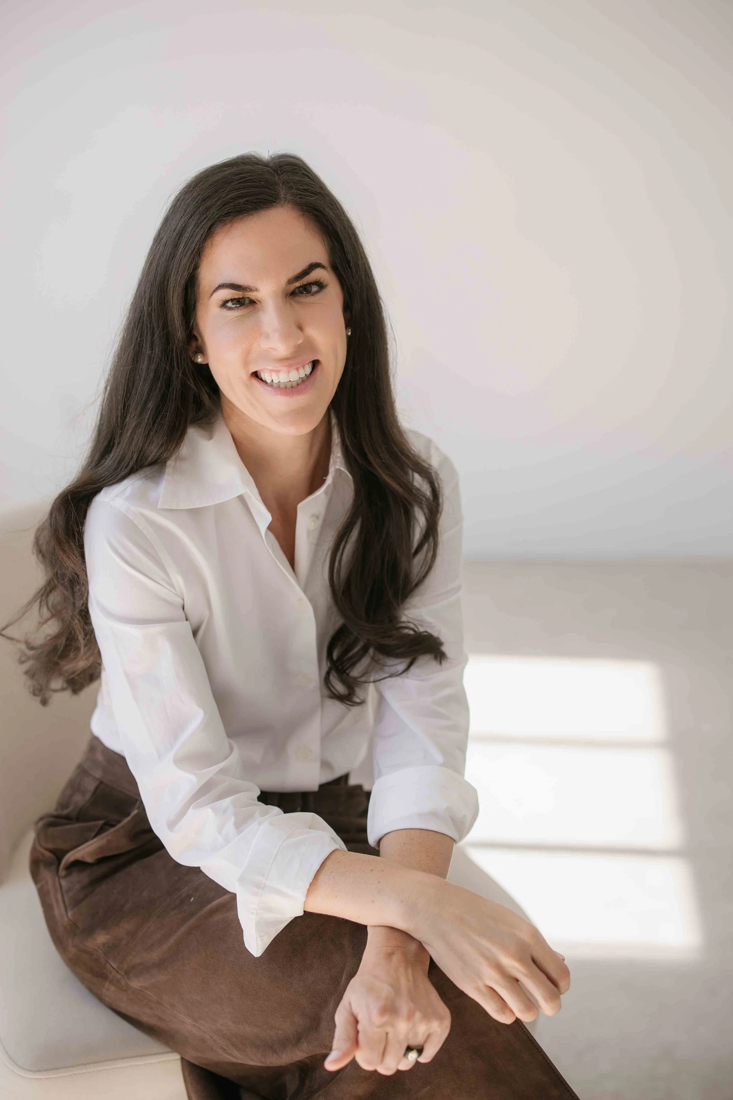 A woman with long dark hair and a white shirt smiling and sitting on a beige chair.