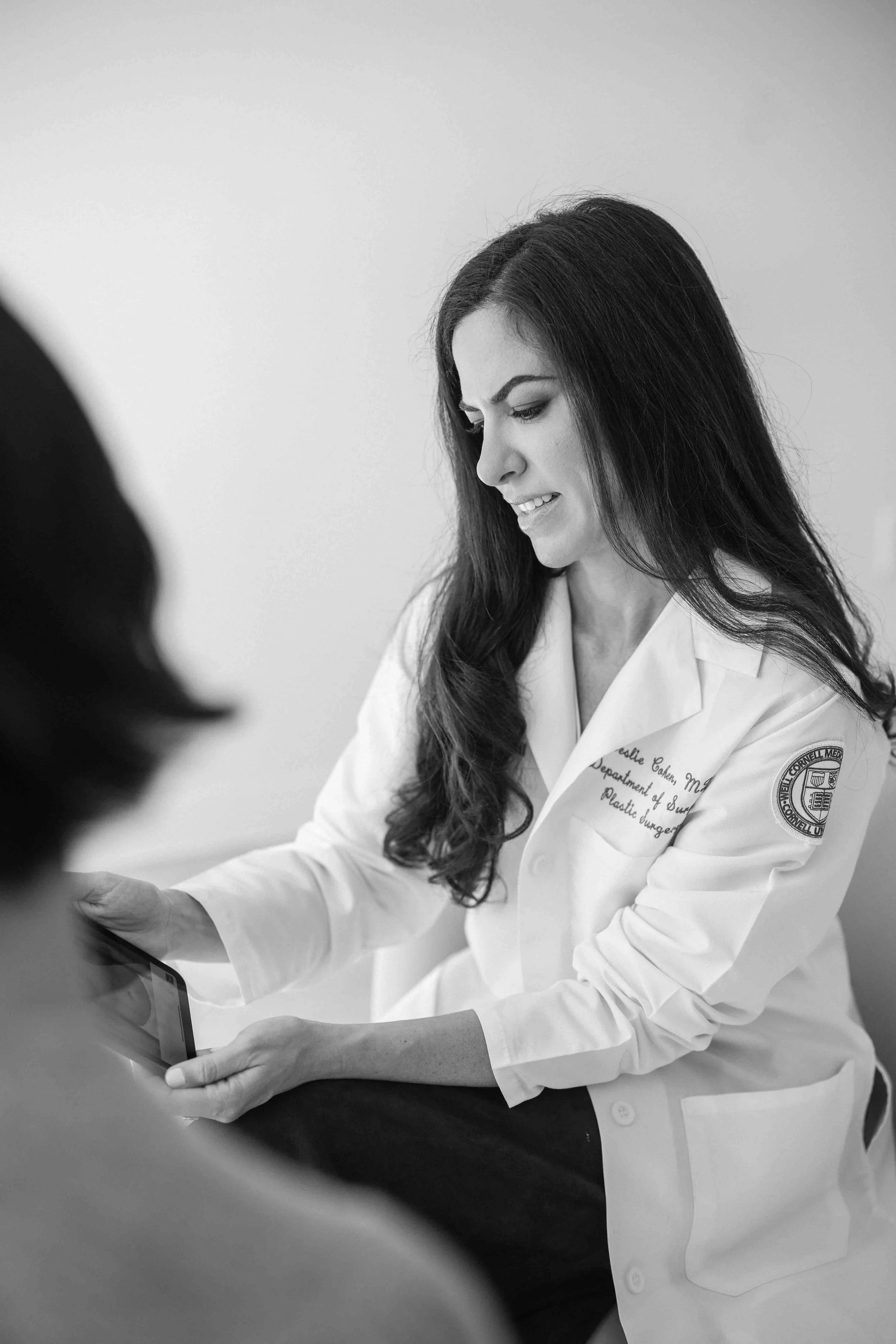 Black and white photo of a woman in a medical coat looking at a tablet, with a blurred person in the foreground.