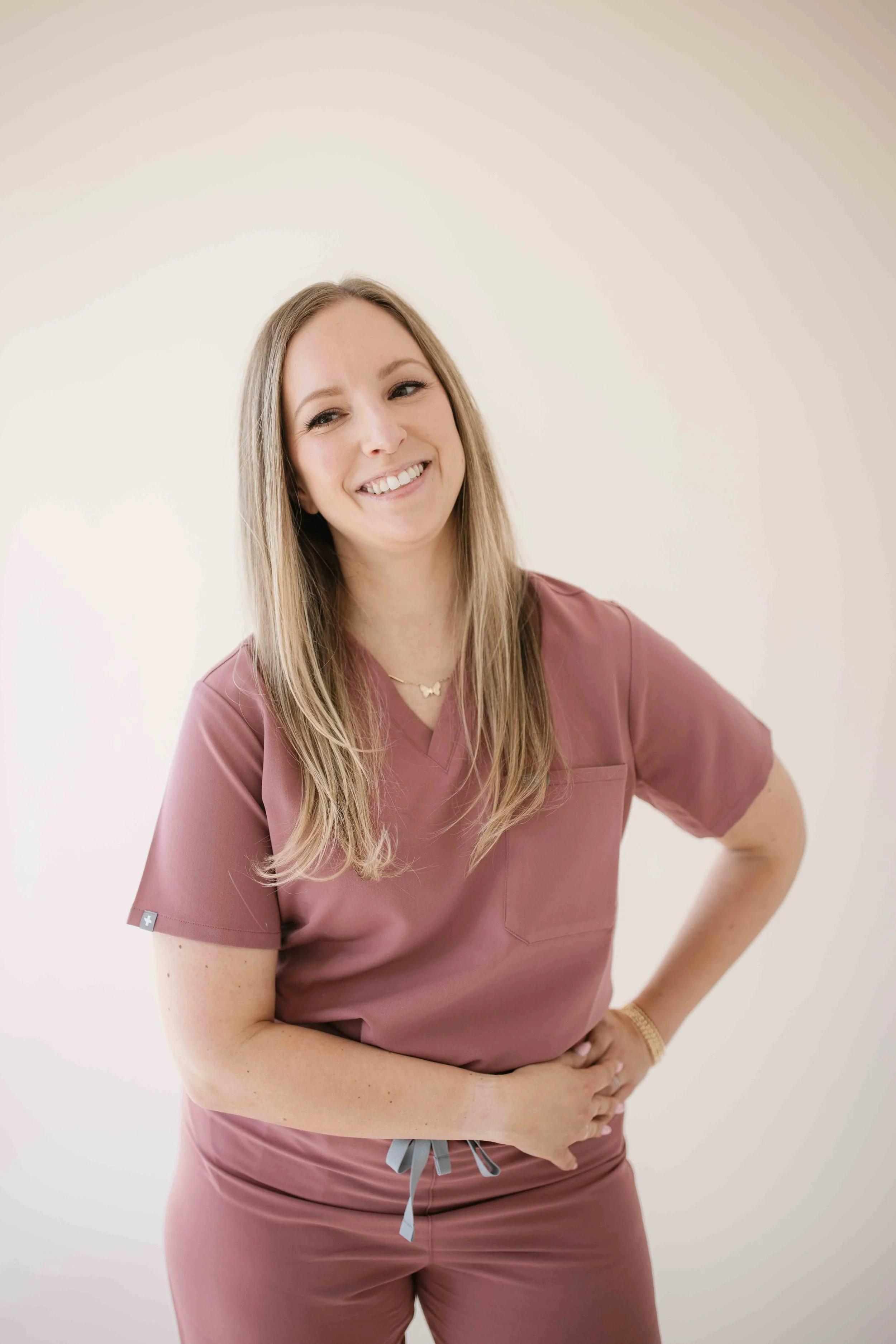 A smiling woman in pink medical scrubs standing with one hand on her hip against a plain white background.