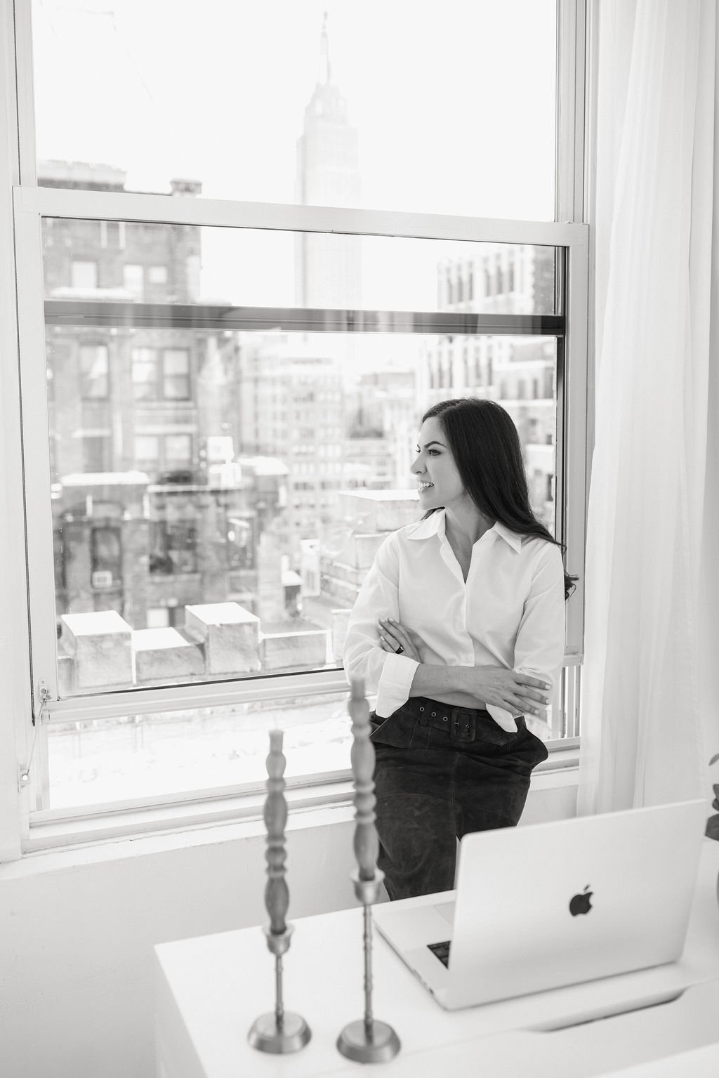 A woman with long dark hair, wearing a white shirt, standing and looking out a window in an urban office setting with skyscrapers in the background.