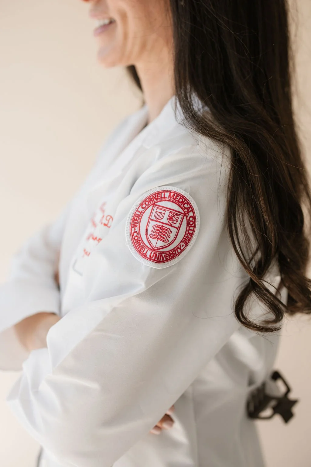 Close-up of a person wearing a white medical coat with an embroidered red patch on the sleeve, indicating affiliation with The Cornell Medical College at Cornell University. The person has long, dark, wavy hair.