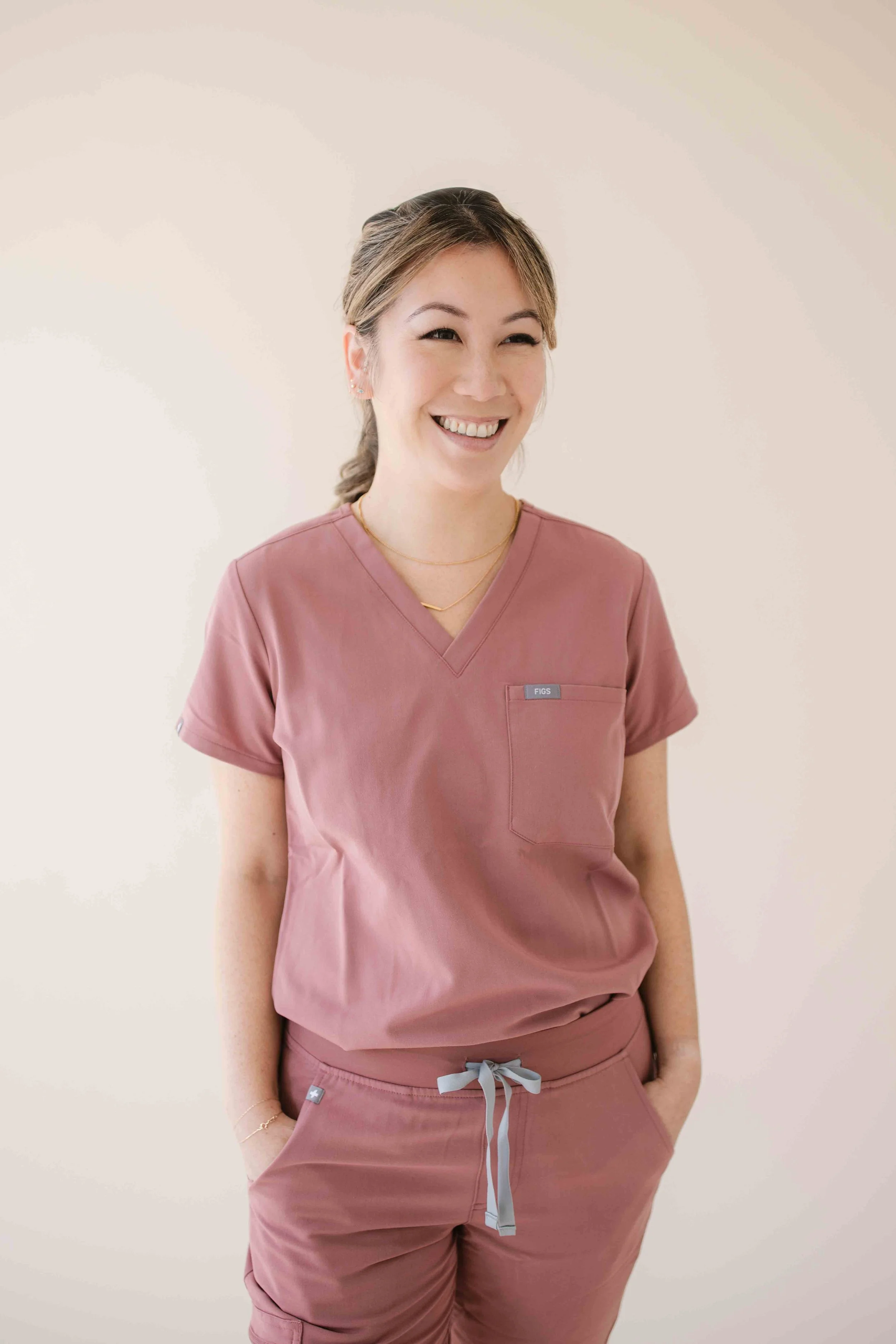 A woman wearing pink scrubs, smiling, with her hands in her pockets, standing against a plain light-colored background.