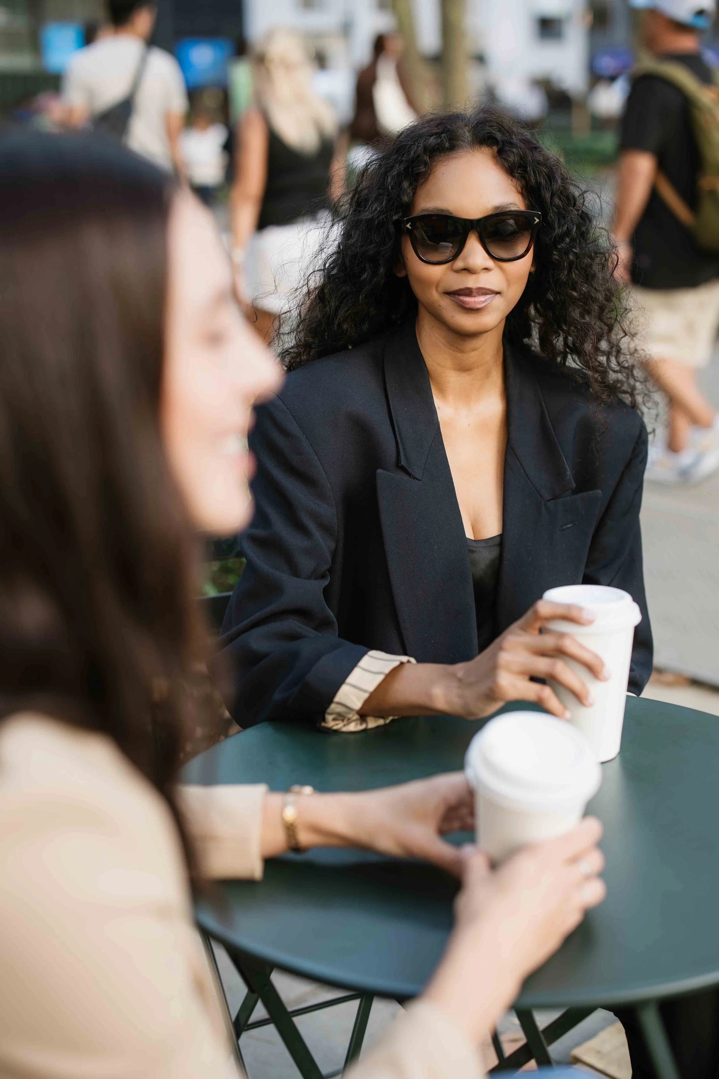Two women sit at a small outdoor table, holding disposable coffee cups. The woman in focus wears sunglasses and a dark blazer, with curly hair. The other woman is blurred in the foreground.