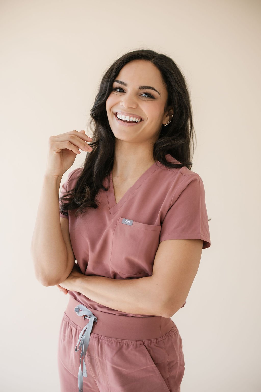 A smiling woman with long dark hair wearing pink medical scrubs, standing against a plain light-colored background.