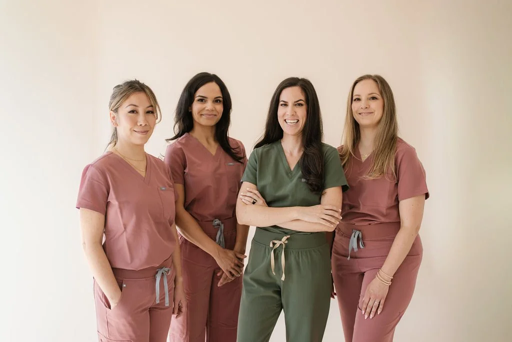 Four female healthcare professionals wearing scrubs standing together against a plain background, smiling and posing for the photo.