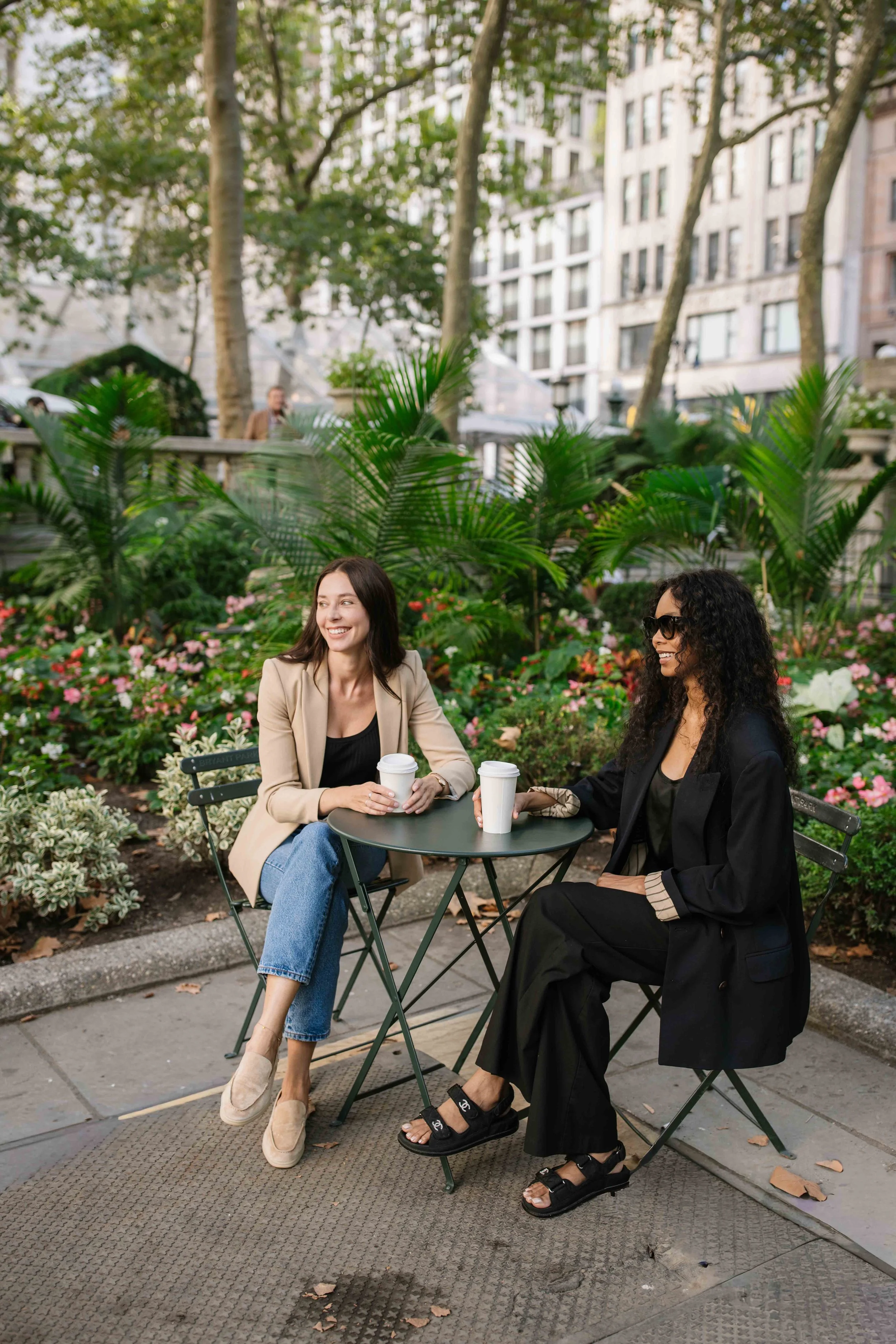 Two women sitting at a small outdoor table in a park, smiling and holding coffee cups, with lush greenery and flowers around them.