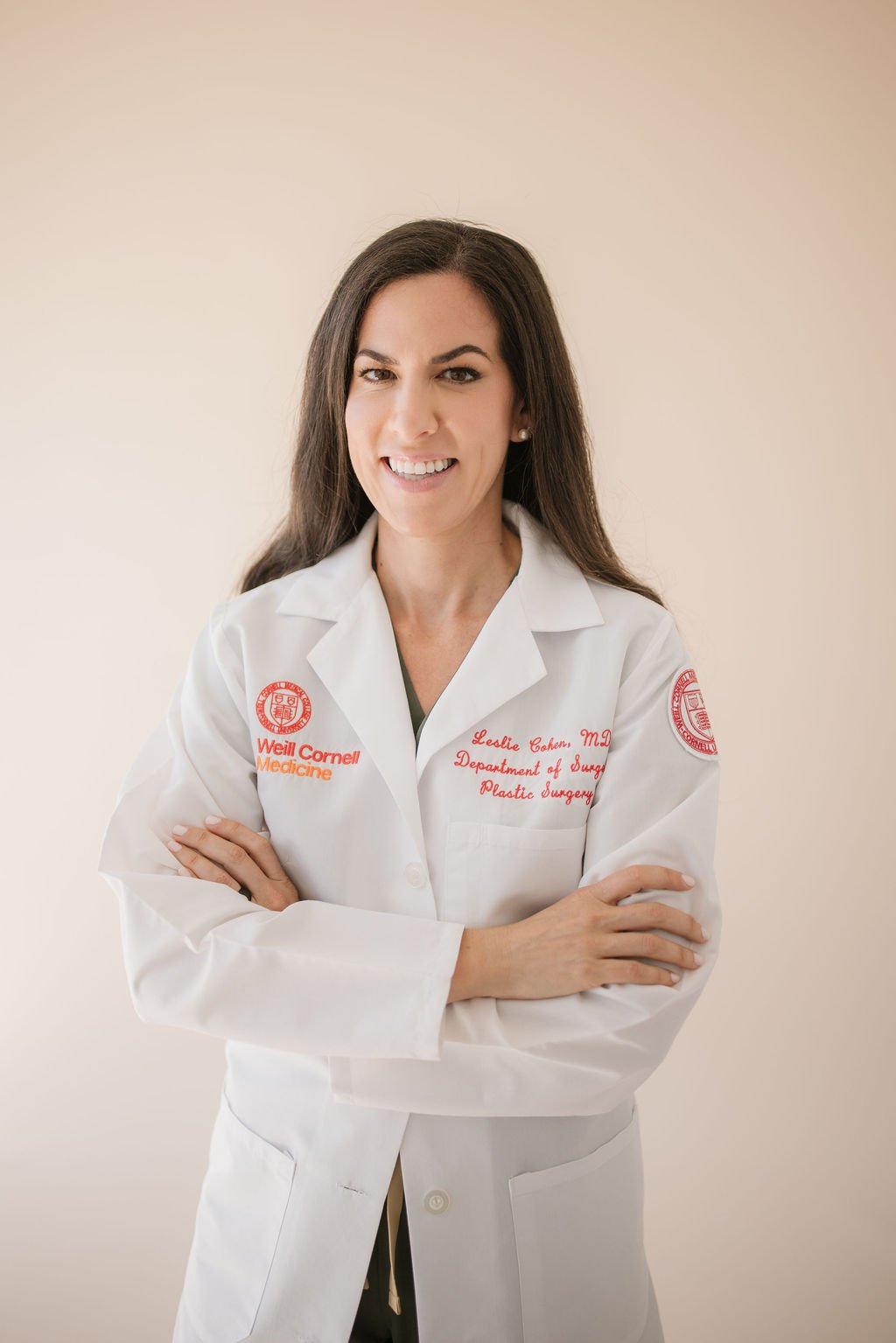 A female doctor in a white coat with embroidered text, smiling and crossing her arms, standing against a beige background.