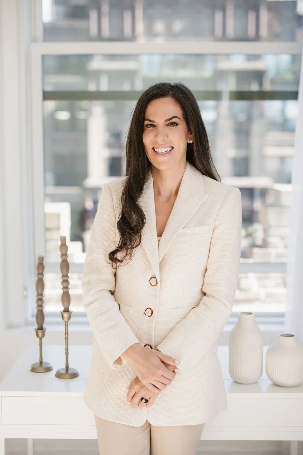 A woman with long dark hair wearing a cream-colored suit, smiling and standing indoors with a city view in the background.