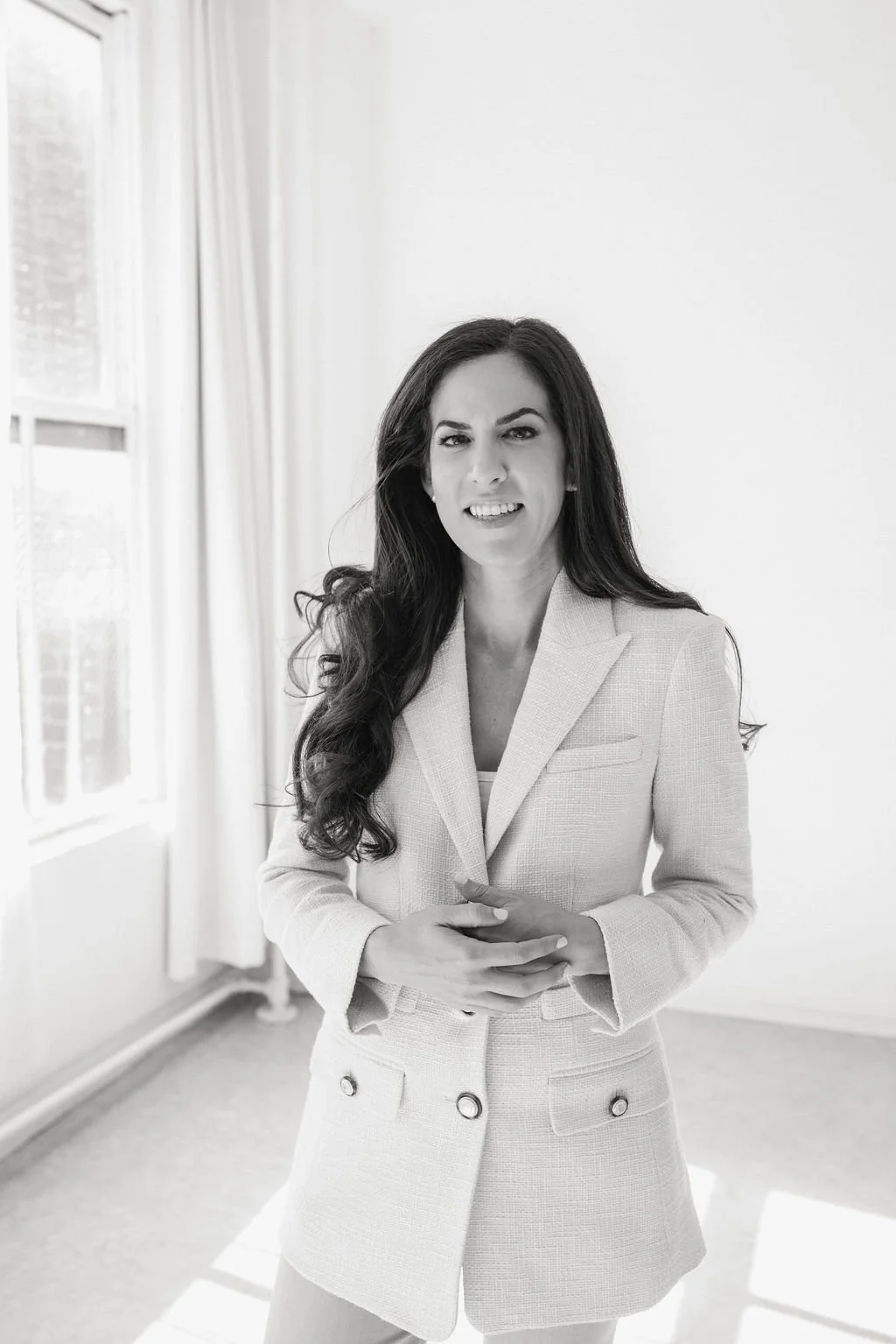 Black and white photo of a woman with long dark hair in a blazer, standing indoors near a window, smiling slightly and looking at the camera.