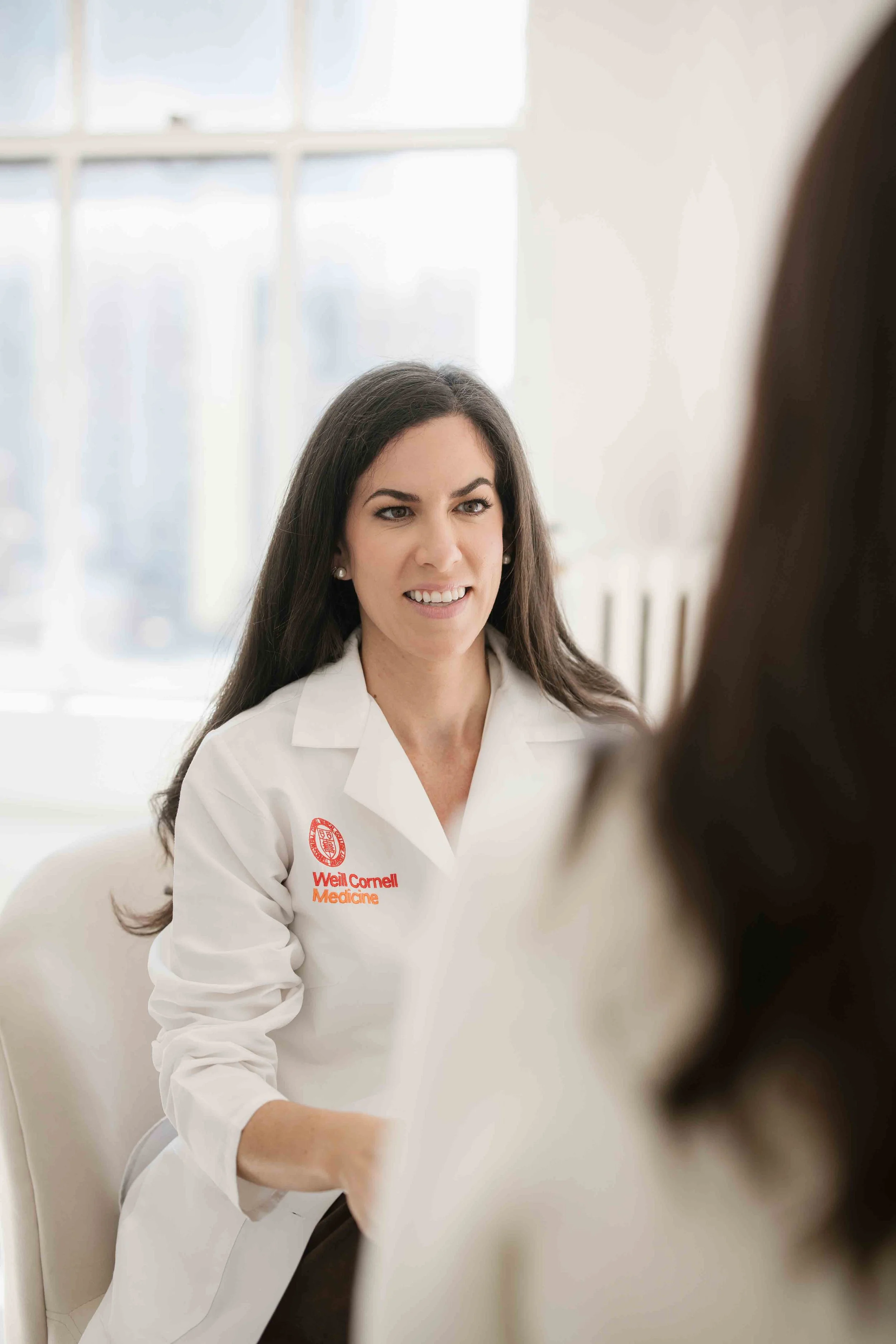 A woman in a white lab coat with 'Weill Cornell Medicine' embroidered on it, sitting and facing a person with long hair, in a well-lit room with large windows.