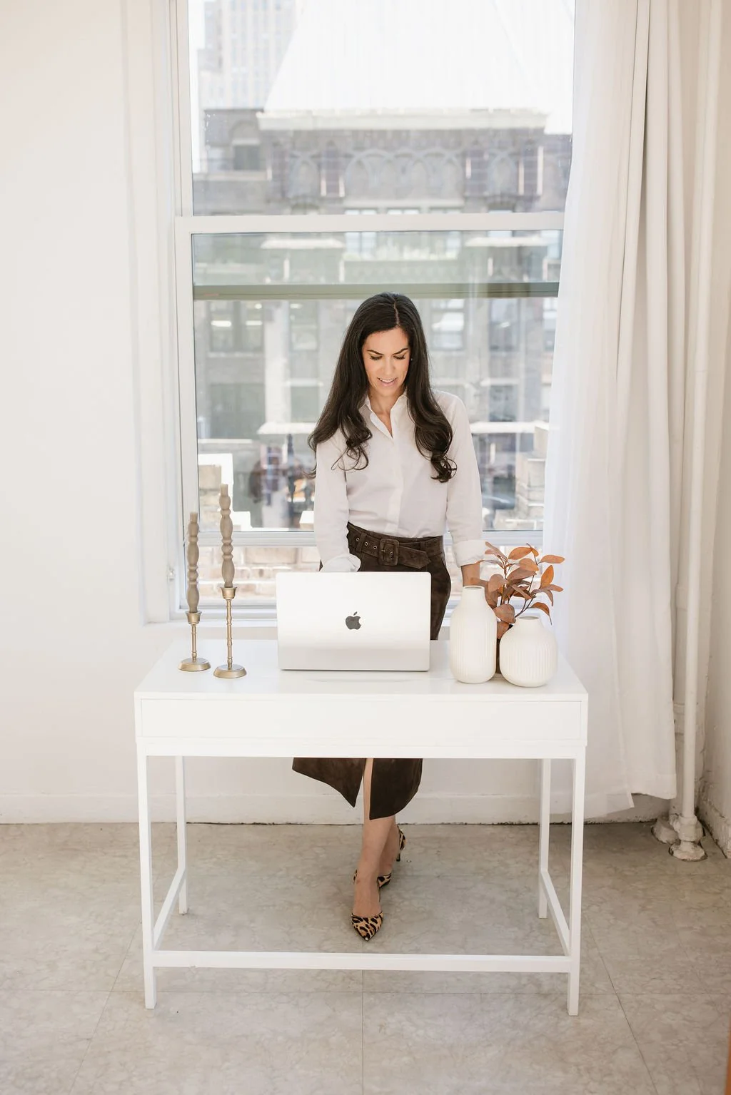 Woman standing behind a white desk with a silver MacBook, two vases with a plant, and three gold candlesticks in front of a large window with a city view.