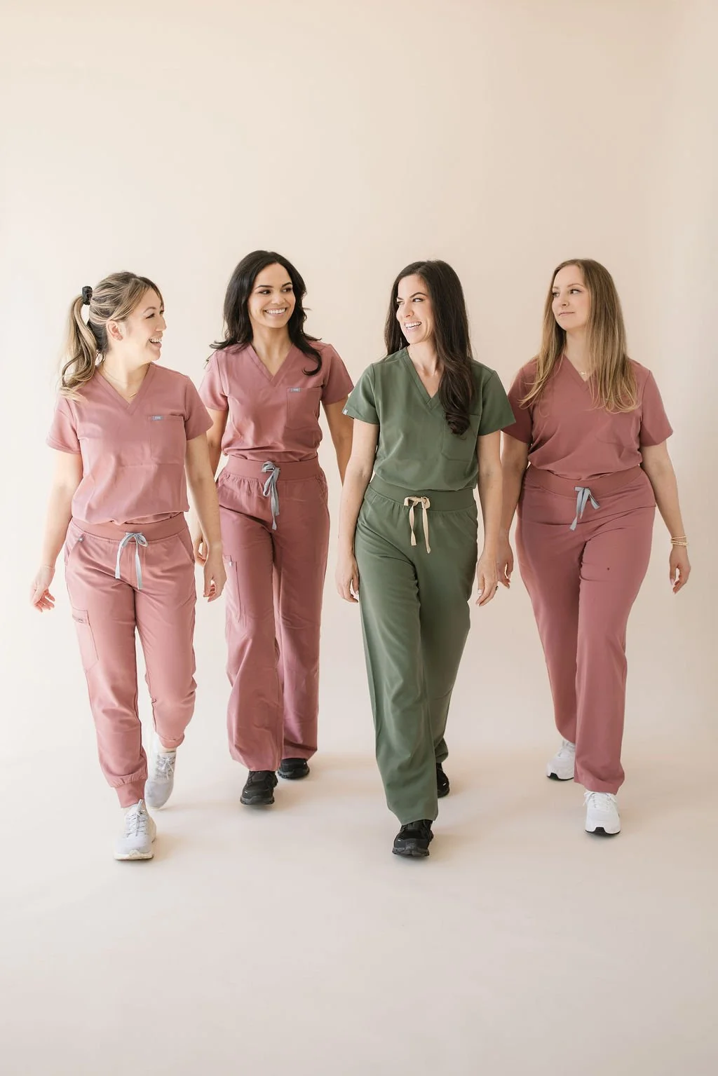 Four women wearing medical scrubs walking together and smiling in a neutral indoor space.