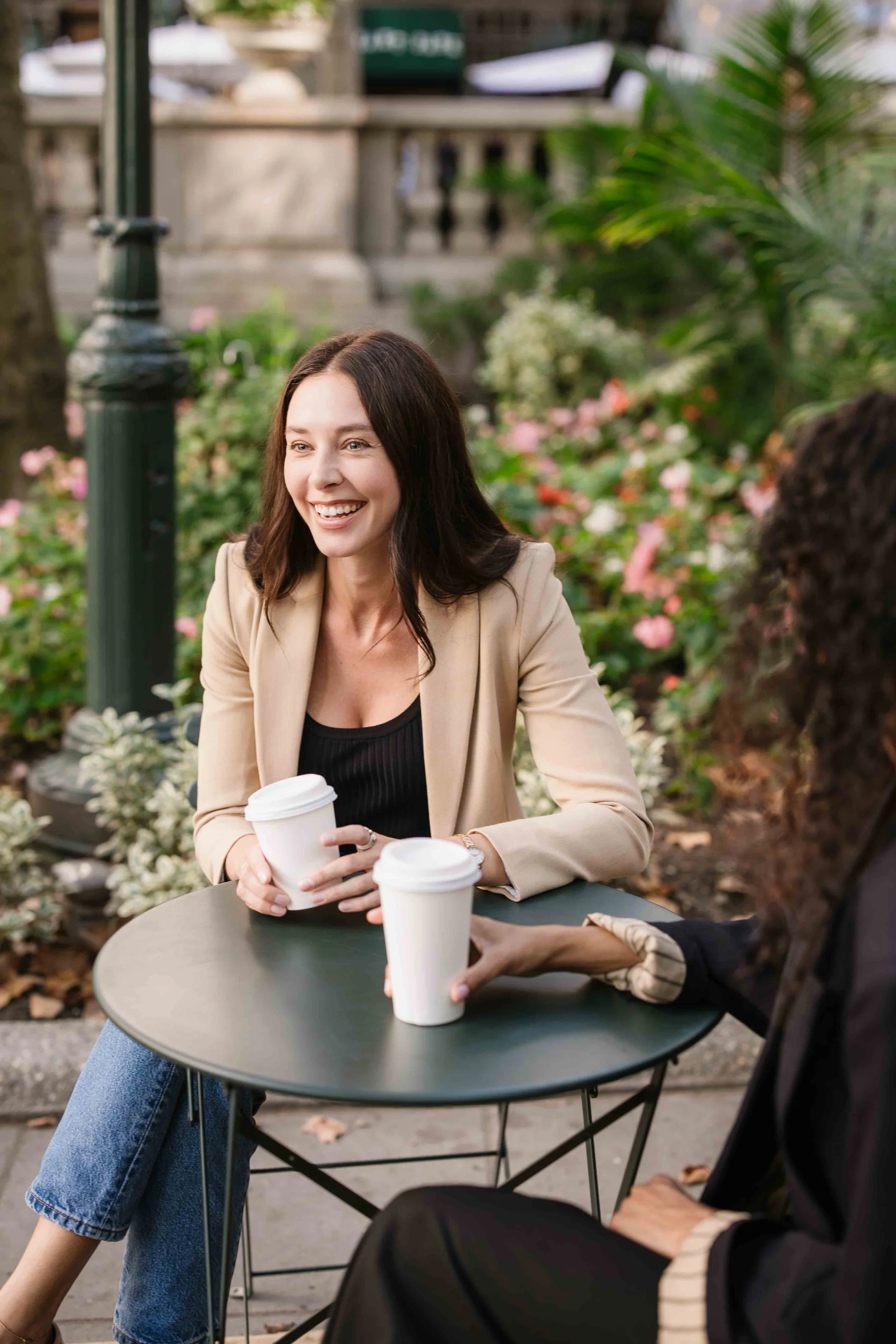 Two women sitting at a small outdoor table with takeaway coffee cups, smiling and talking, surrounded by greenery and flowers.