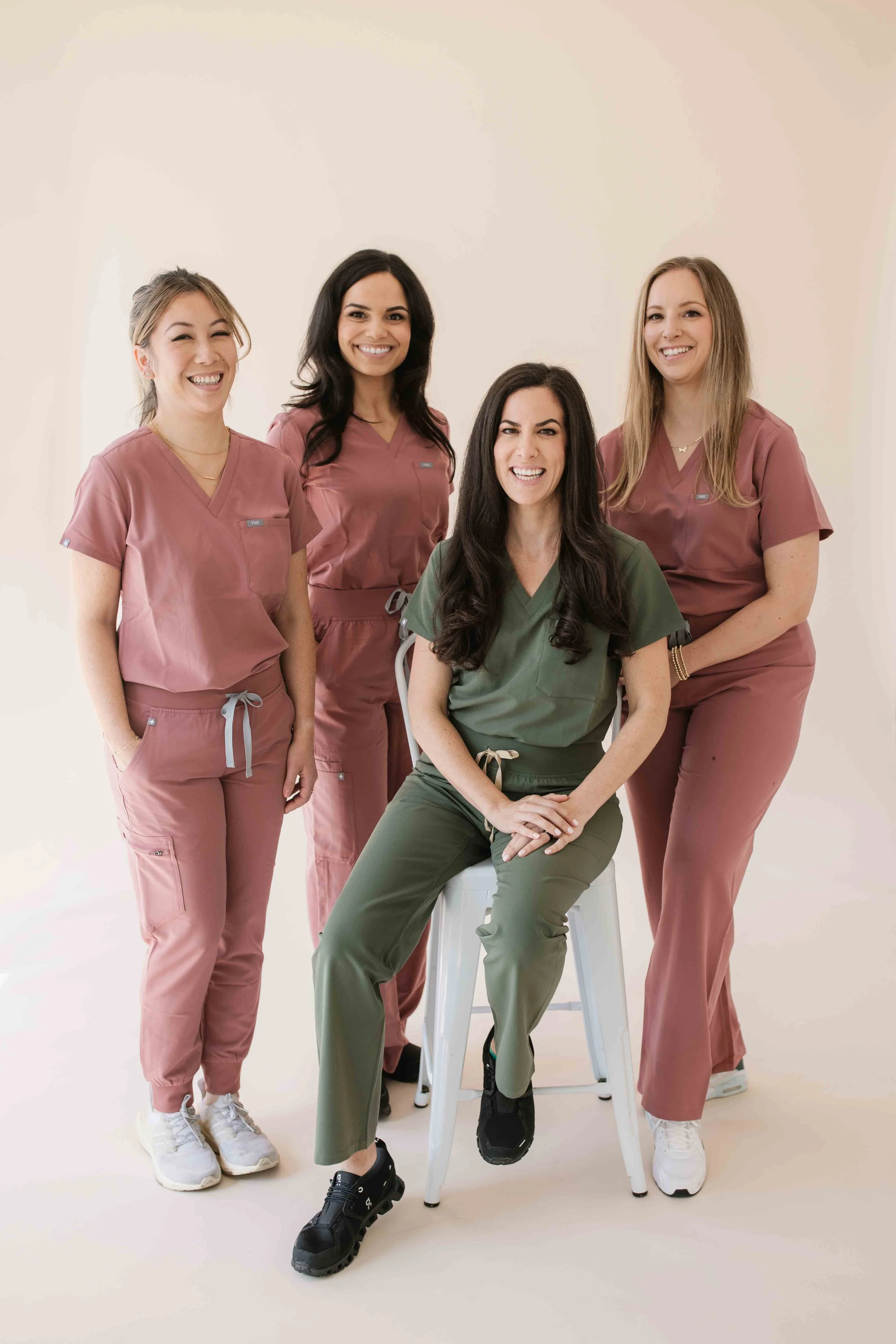 A group of four women medical professionals, three standing and one seated on a stool, wearing scrubs of pink and green, smiling at the camera against a neutral background.