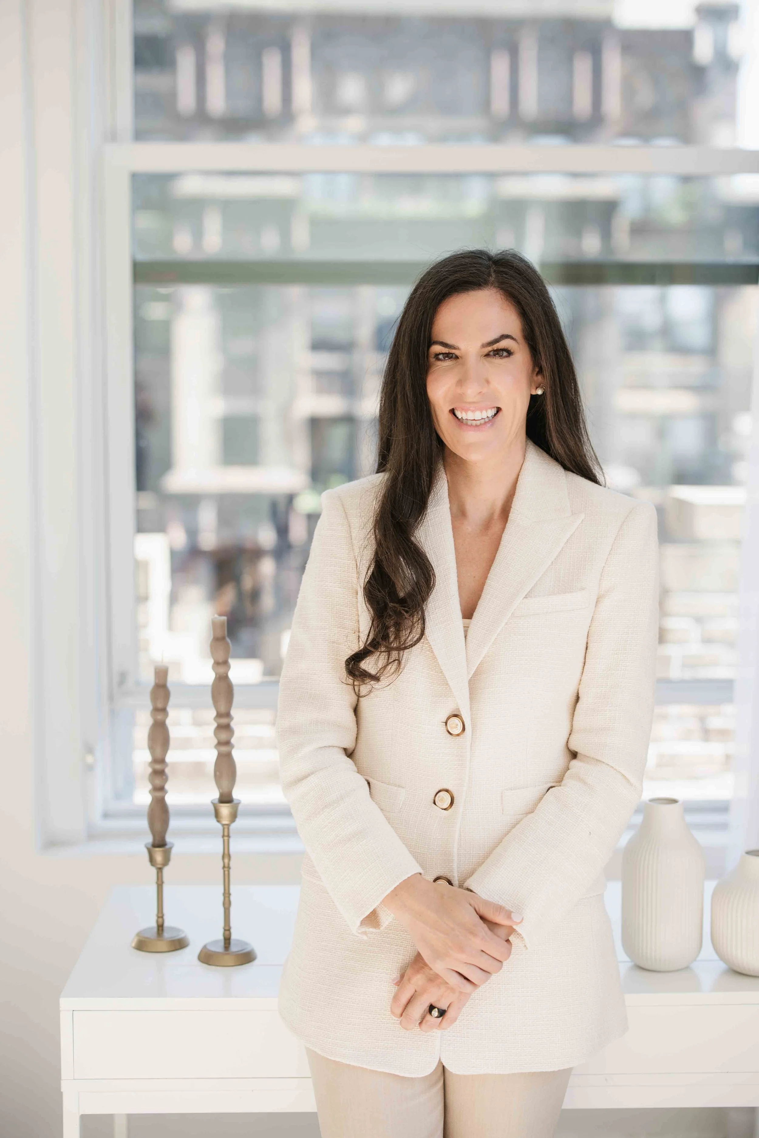 A woman standing indoors, smiling at the camera, dressed in a light-colored blazer and pants, with candles and vases on a table behind her, and a window in the background.